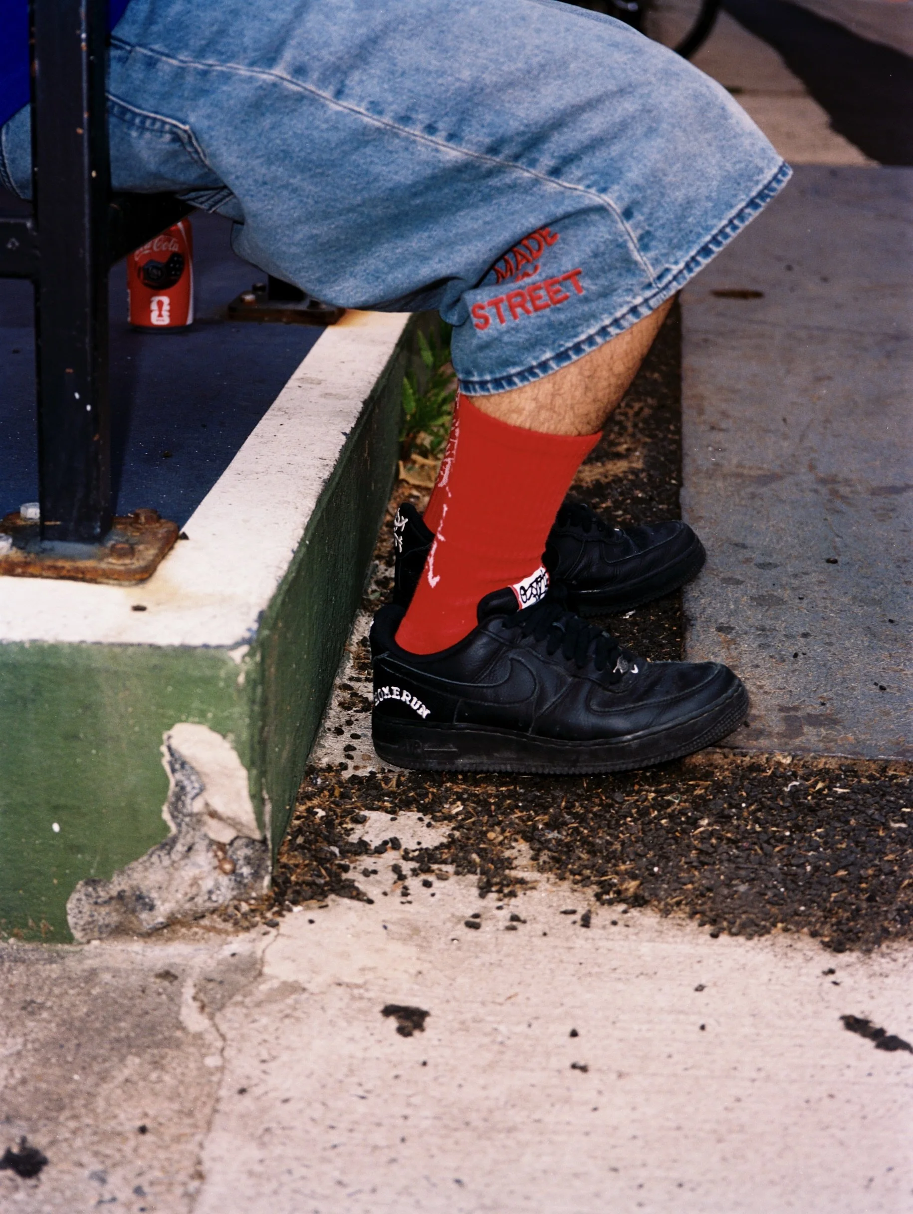 Close-up of a person's lower body sitting on the curb, wearing blue denim shorts with red embroidered text and bright red socks, with black sneakers, near a parking lot with scattered dirt and debris.