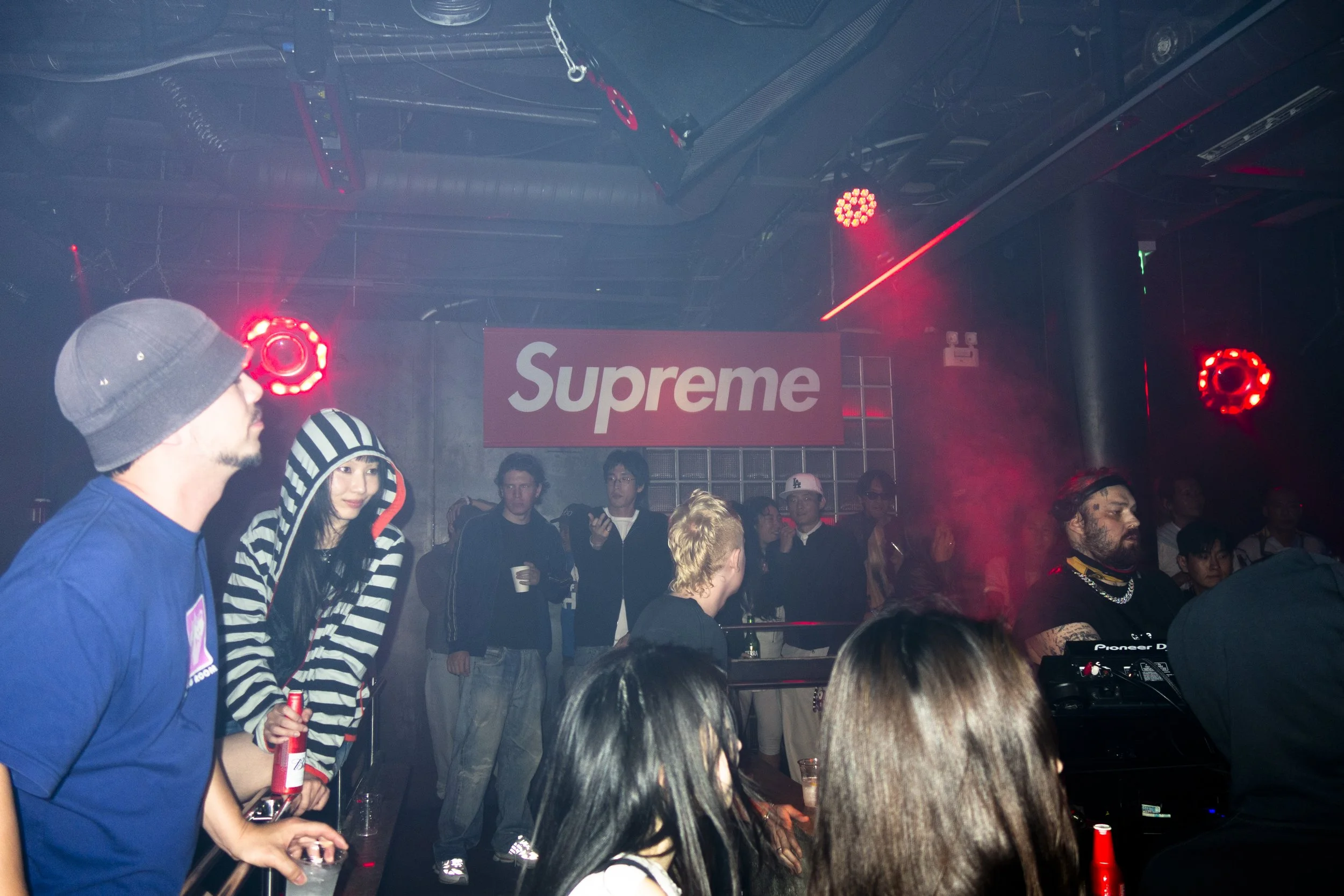 People dancing and socializing in a nightclub with red lighting and a prominent "Supreme" sign on the back wall.