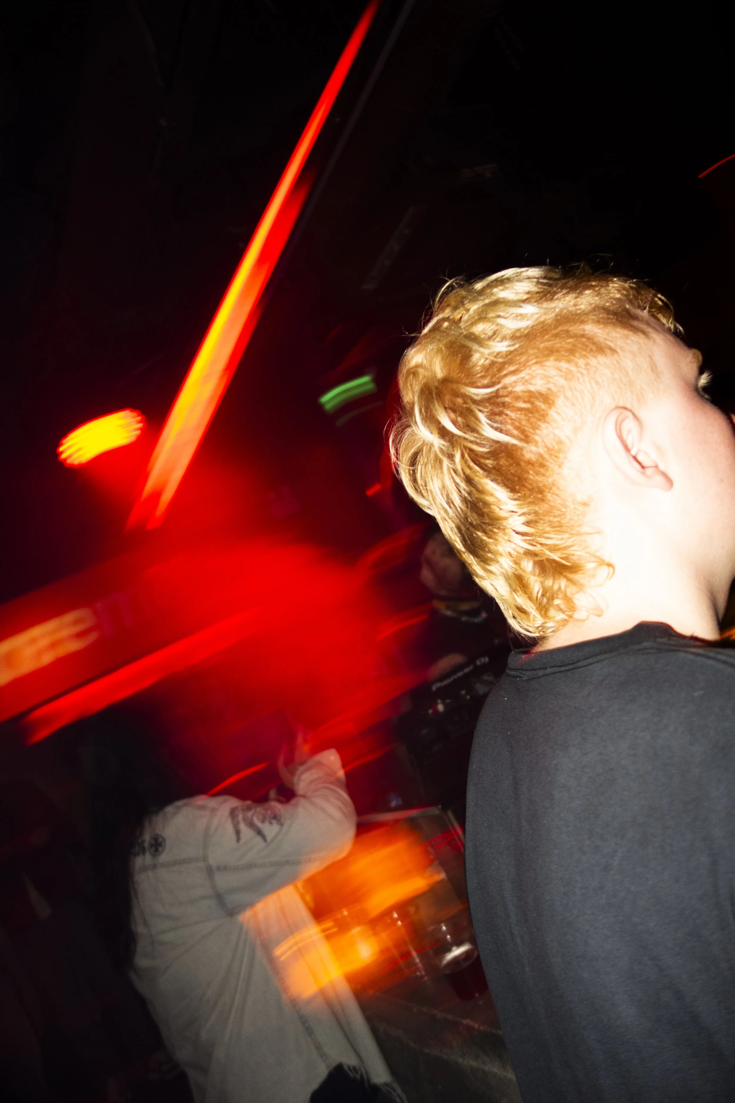 A person with blond hair facing to the right in a dark environment with red and yellow lights and motion blur.