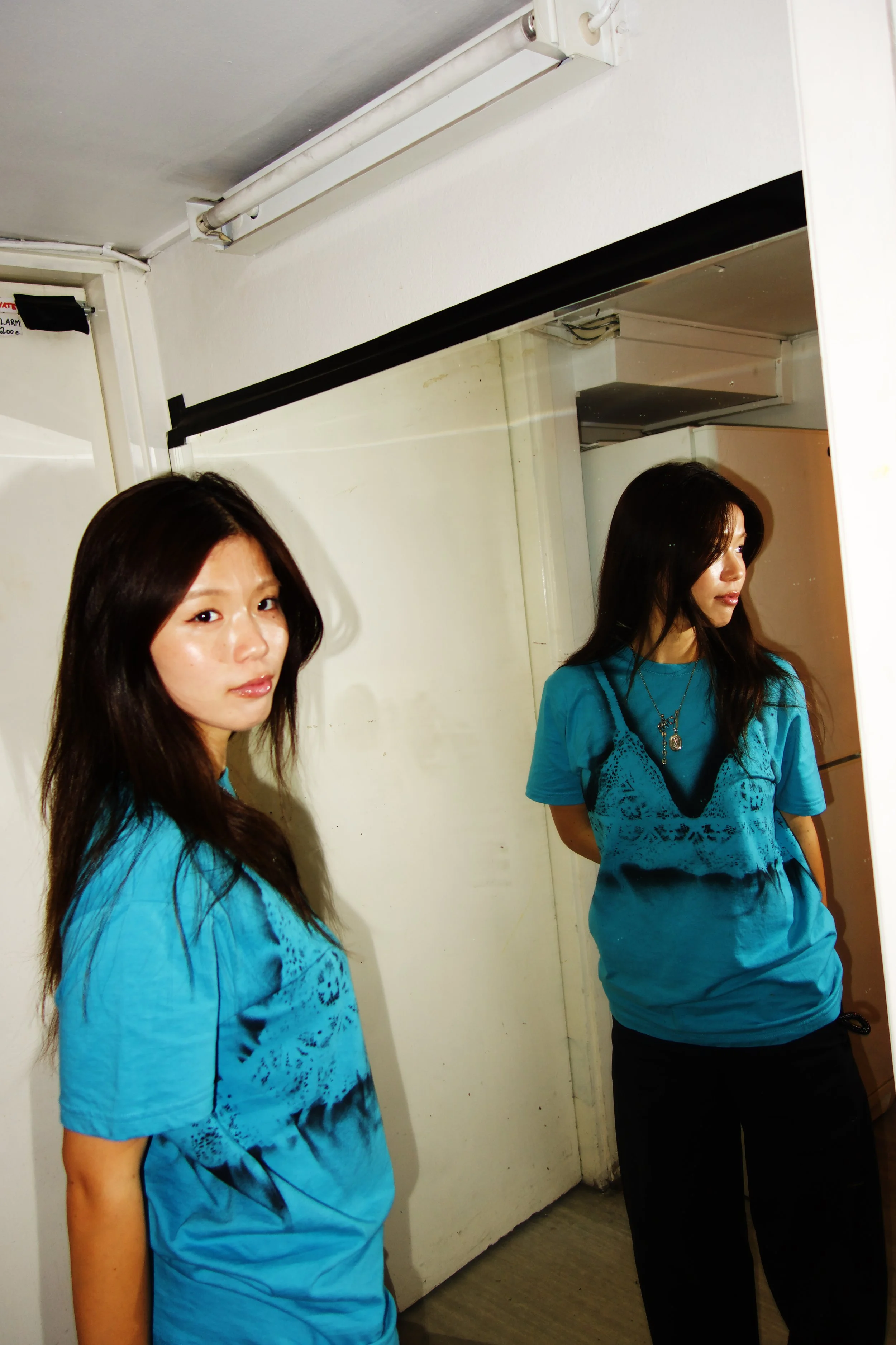 Two women with long dark hair wearing turquoise shirts with black design, standing in a room with white walls and a door. One woman is looking at the camera, and the other is looking to the side.