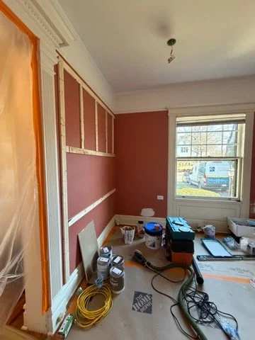 A home under construction. A wood frame is shown on a dark pink wall. Tools and paint are on the ground