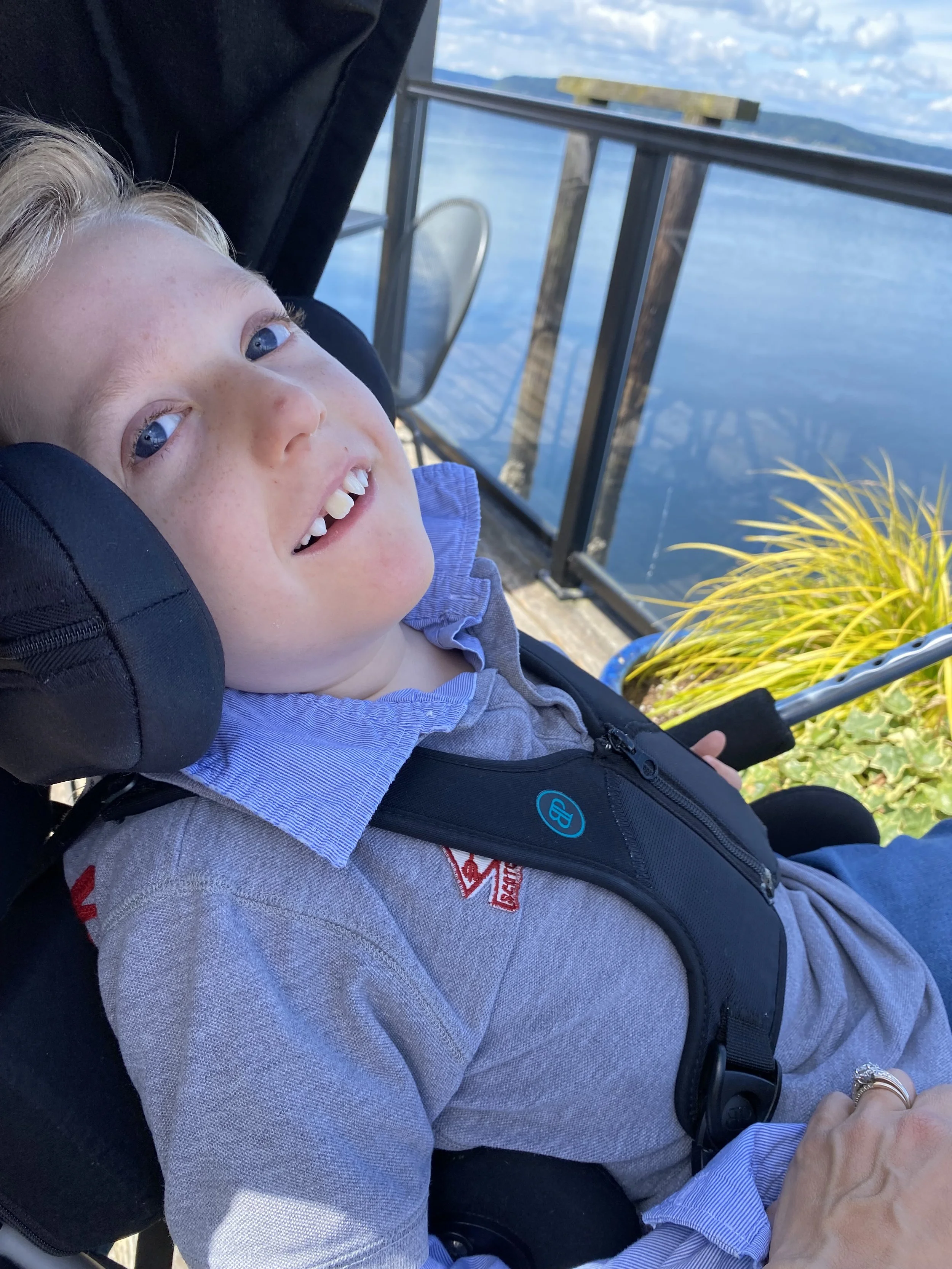 Jack Henry Wilkes smiles. He is in a wheelchair, looking at the camera, enjoying a warm day with his mom.