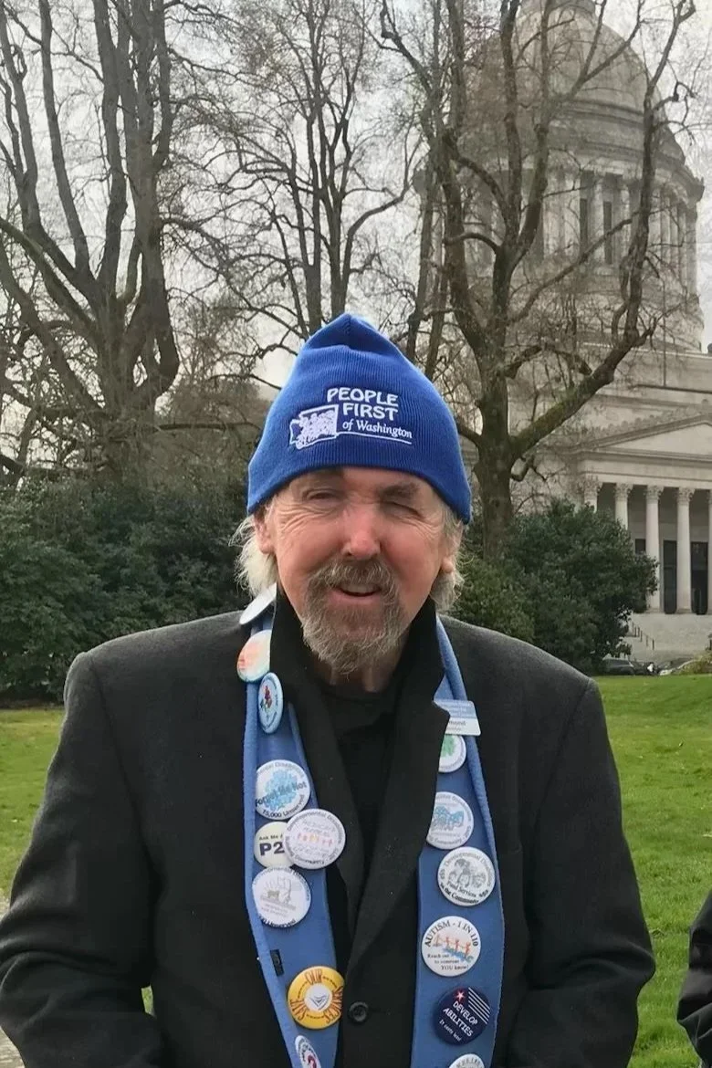 Picture of Mike Raymond wearing a blue People First of Washington hat, standing in front of the Olympia Capitol building.
