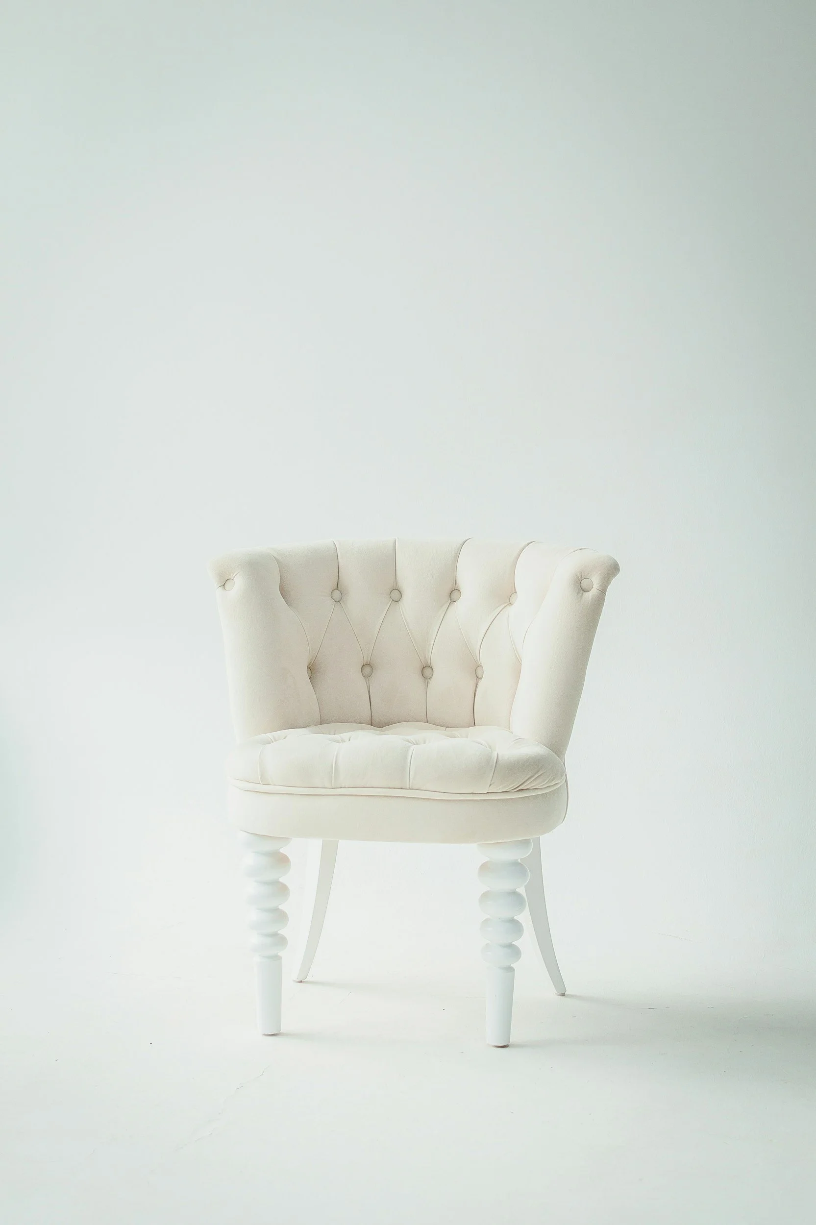 White upholstered chair with button tufting and turned legs sitting on a white floor against a white wall.