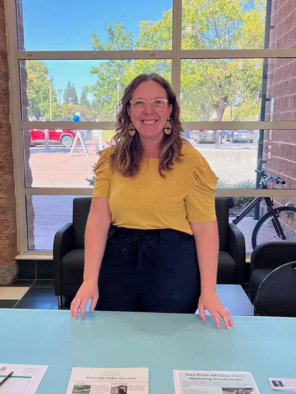 A woman standing behind a table with papers, smiling indoors with a large window showing trees and parked cars outside.