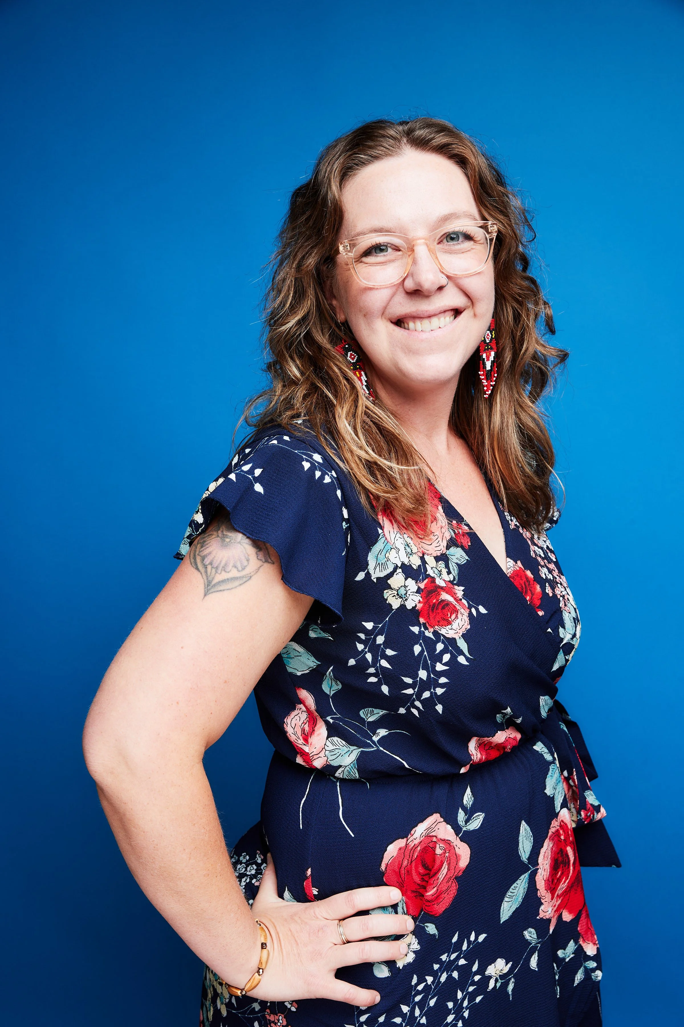 A smiling woman with long wavy brown hair, wearing clear glasses, floral earrings, a floral navy blue dress, standing against a solid blue background.