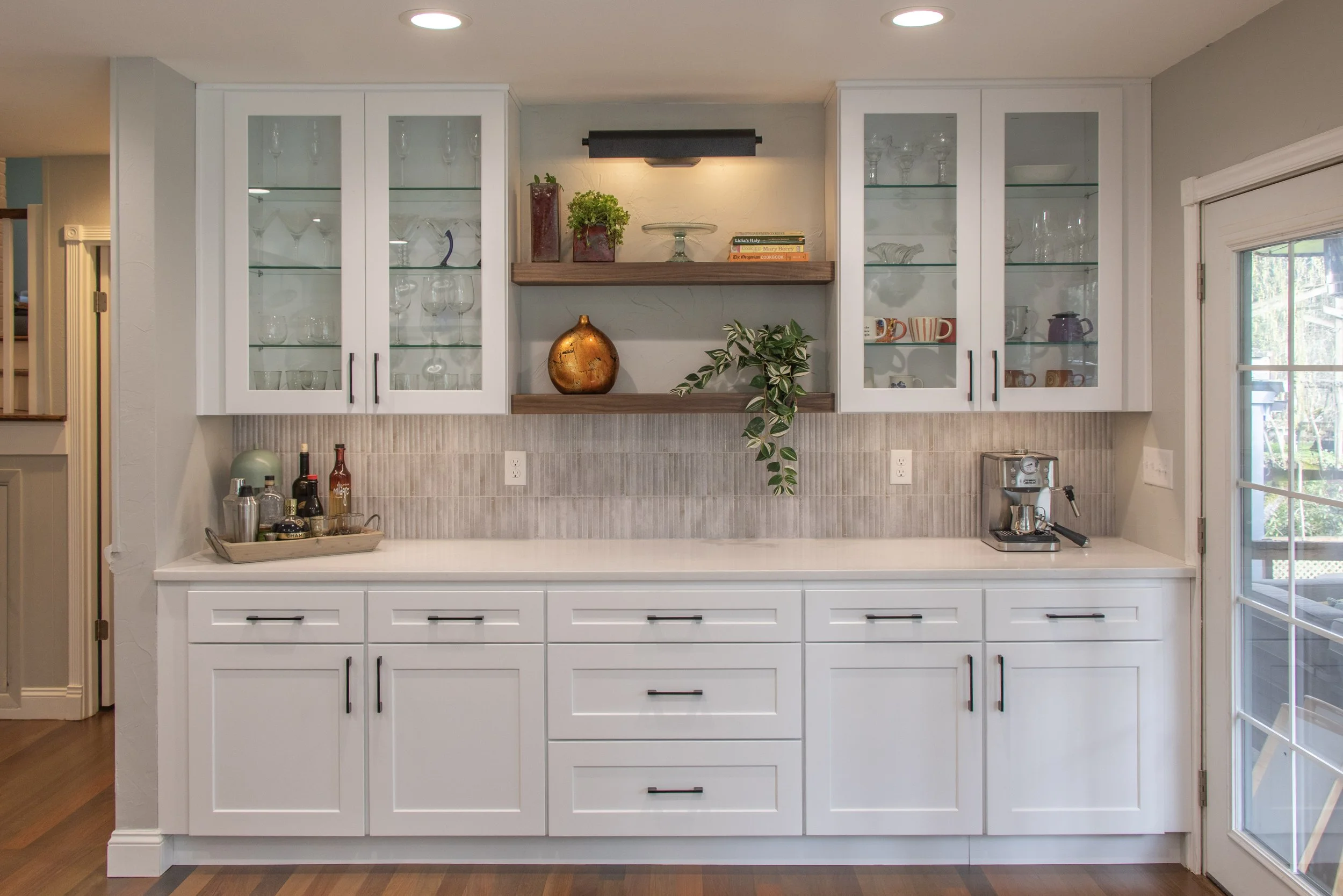 White kitchen cabinets with glass doors, a countertop with bottles, and a section with open shelves holding decorative items and plants, next to a window with a sliding door.