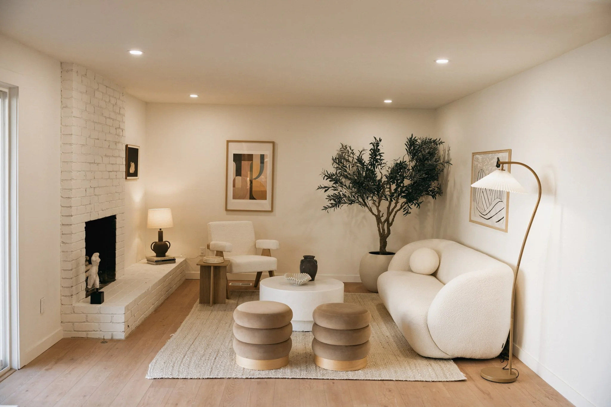 Living room with white walls, a white curved sofa with a round pillow, a large potted tree, abstract artwork, a white armchair, a textured beige area rug, a central white coffee table, two beige ottomans, a standing floor lamp, a table lamp, small de