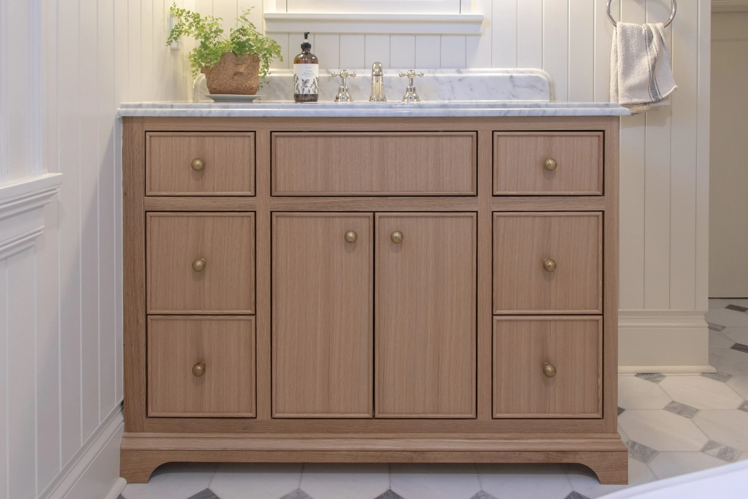 Wooden bathroom vanity with six drawers and brass knobs, white marble countertop, sink, and chrome faucet, in a bathroom with white paneled walls and a towel ring with a towel.