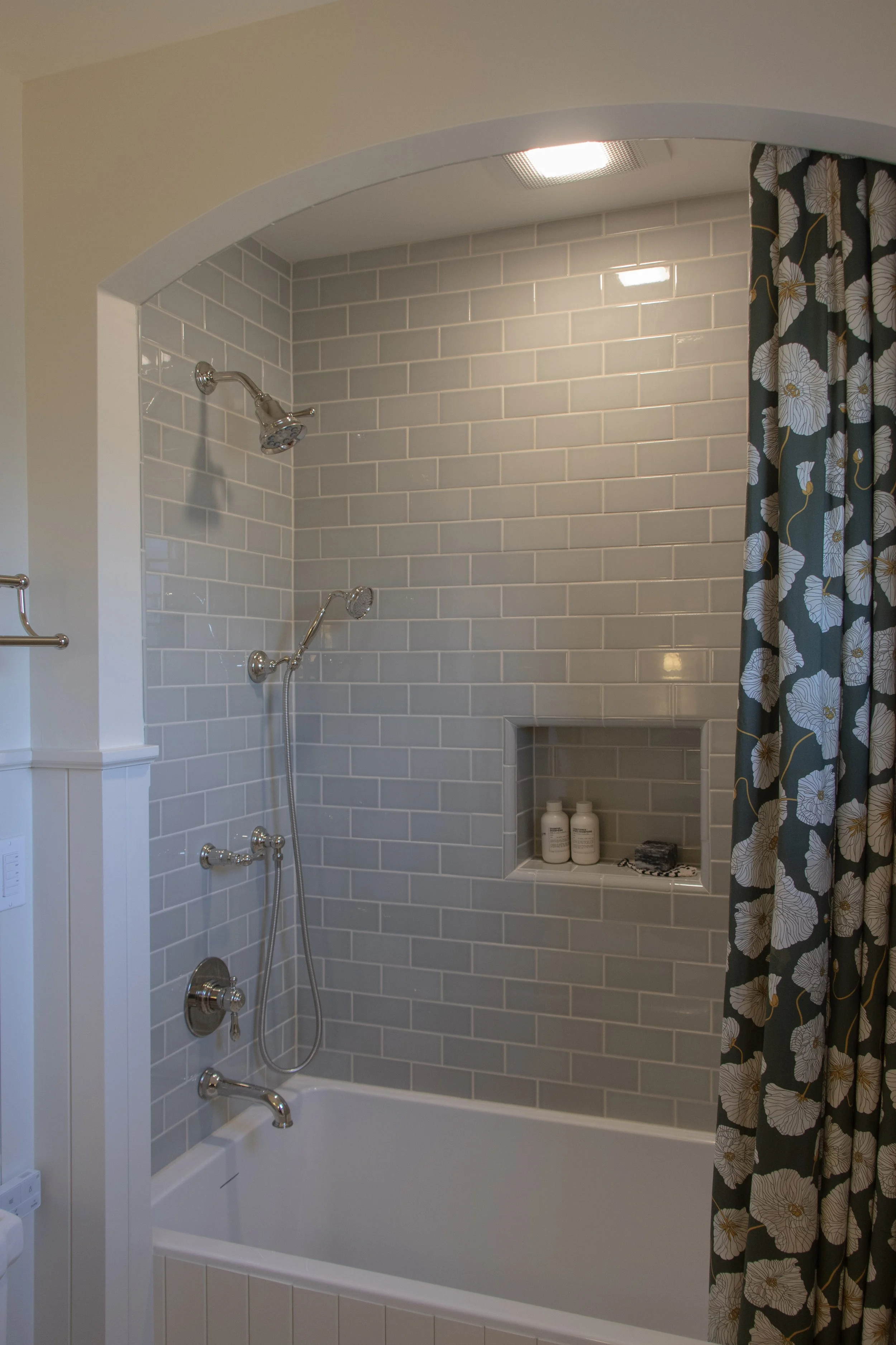 Bathroom with a bathtub and a tiled shower area with a built-in niche holding bath products, a showerhead, and a decorative curtain with floral pattern.
