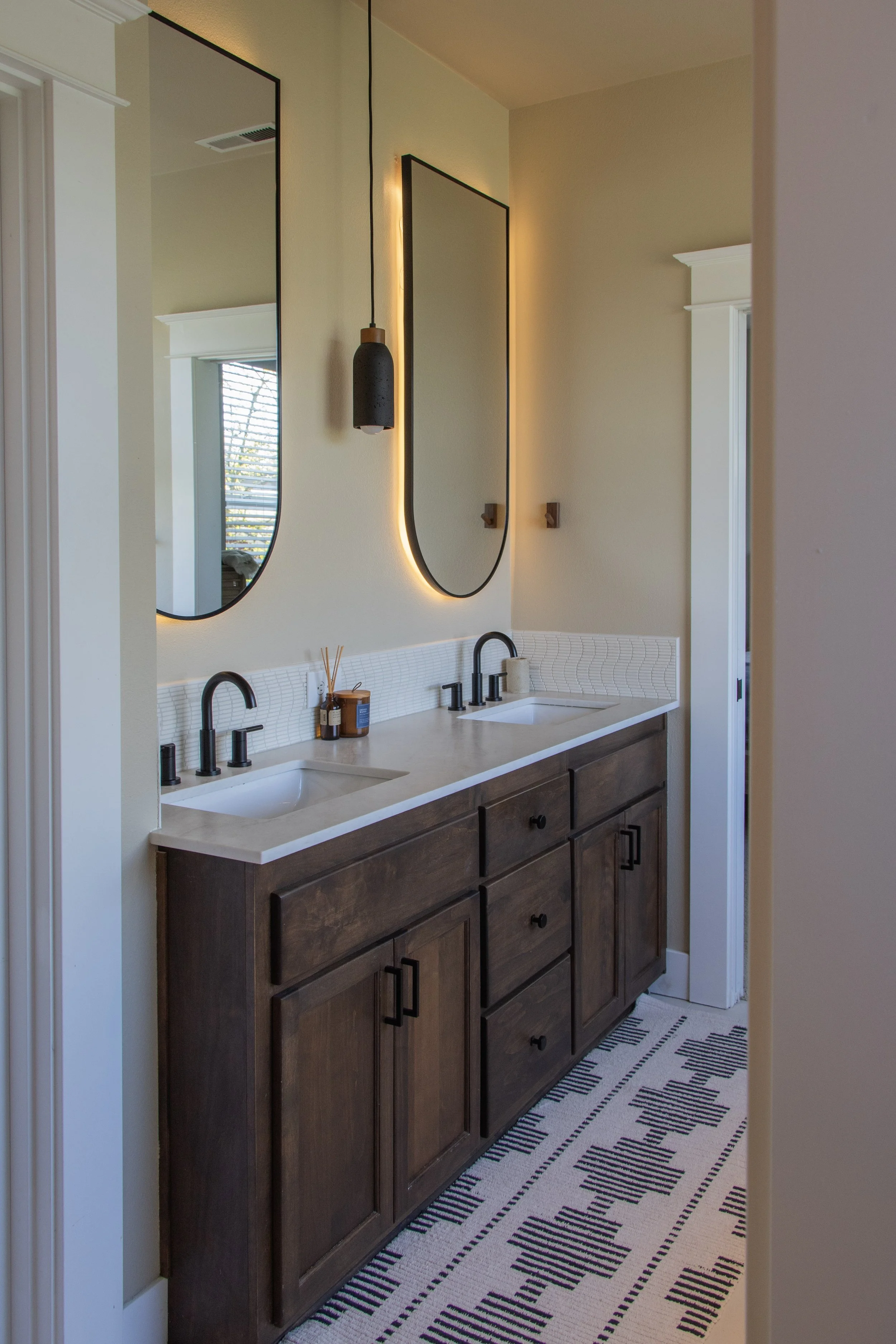 Bathroom vanity with two dark wood cabinets and white countertop, black hardware, two sinks, black faucets, small containers on countertop, framed mirrors with backlit lighting, black pendant light, light-colored walls, patterned black and white rug.