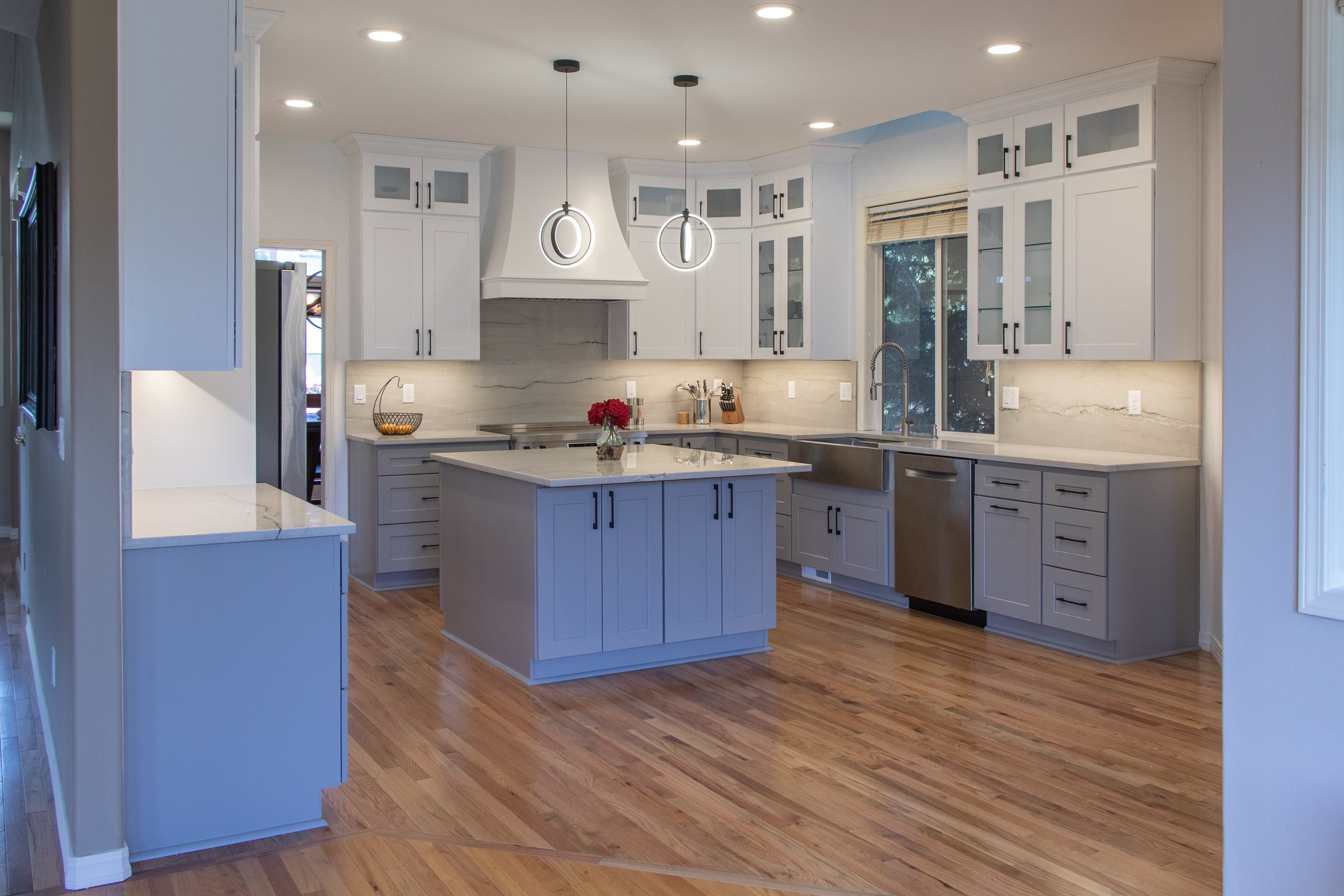 Modern kitchen with white cabinets, marble countertops, hardwood floors, and pendant lighting above a kitchen island. There is a sink beneath a window with a view of trees outside.