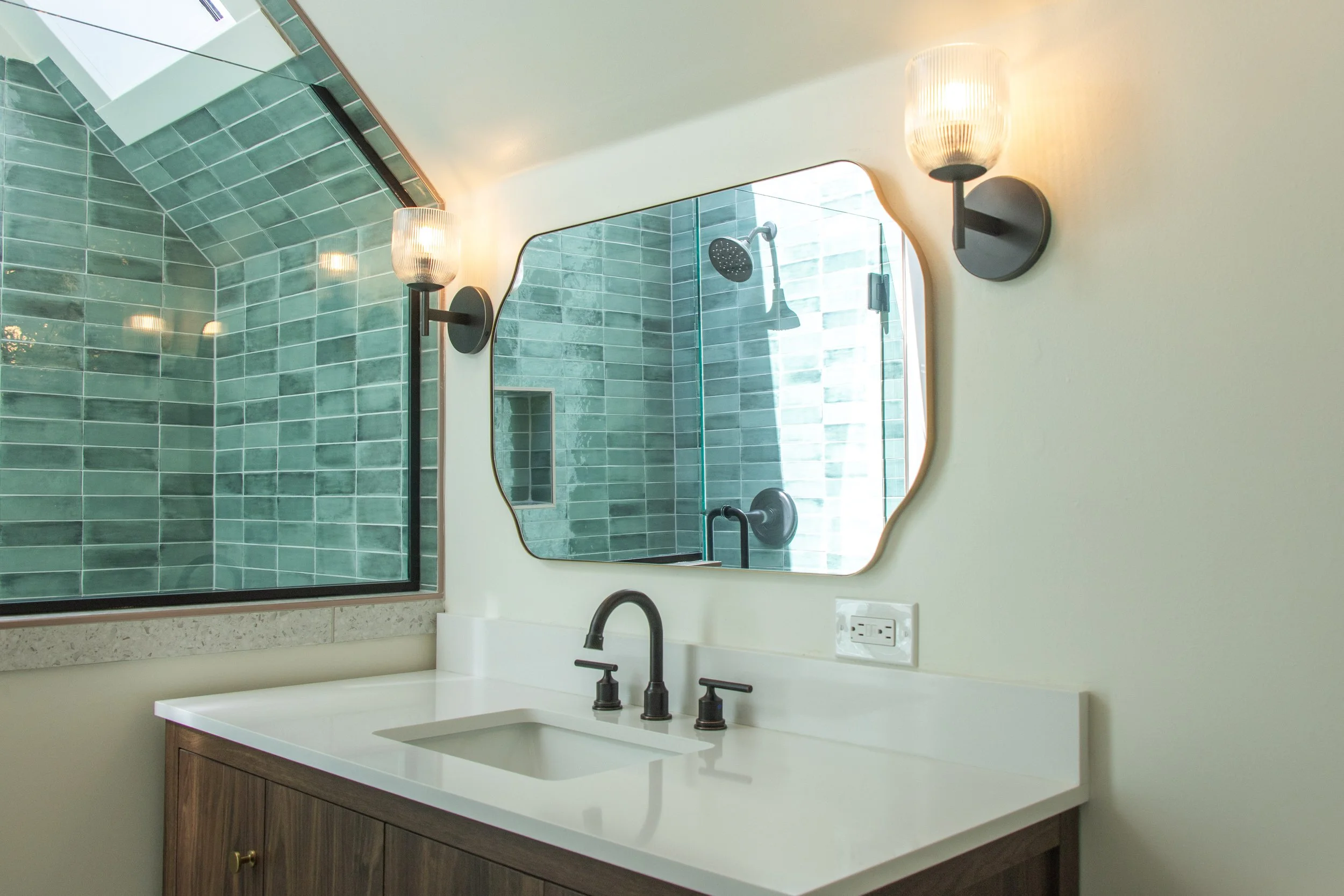 A bathroom vanity with a white countertop, black faucet, and a wooden cabinet. Above, a mirror is mounted on the wall, with a sconce on each side. In the mirror, a green-tiled shower with a round showerhead, a window, and a wall niche are visible.