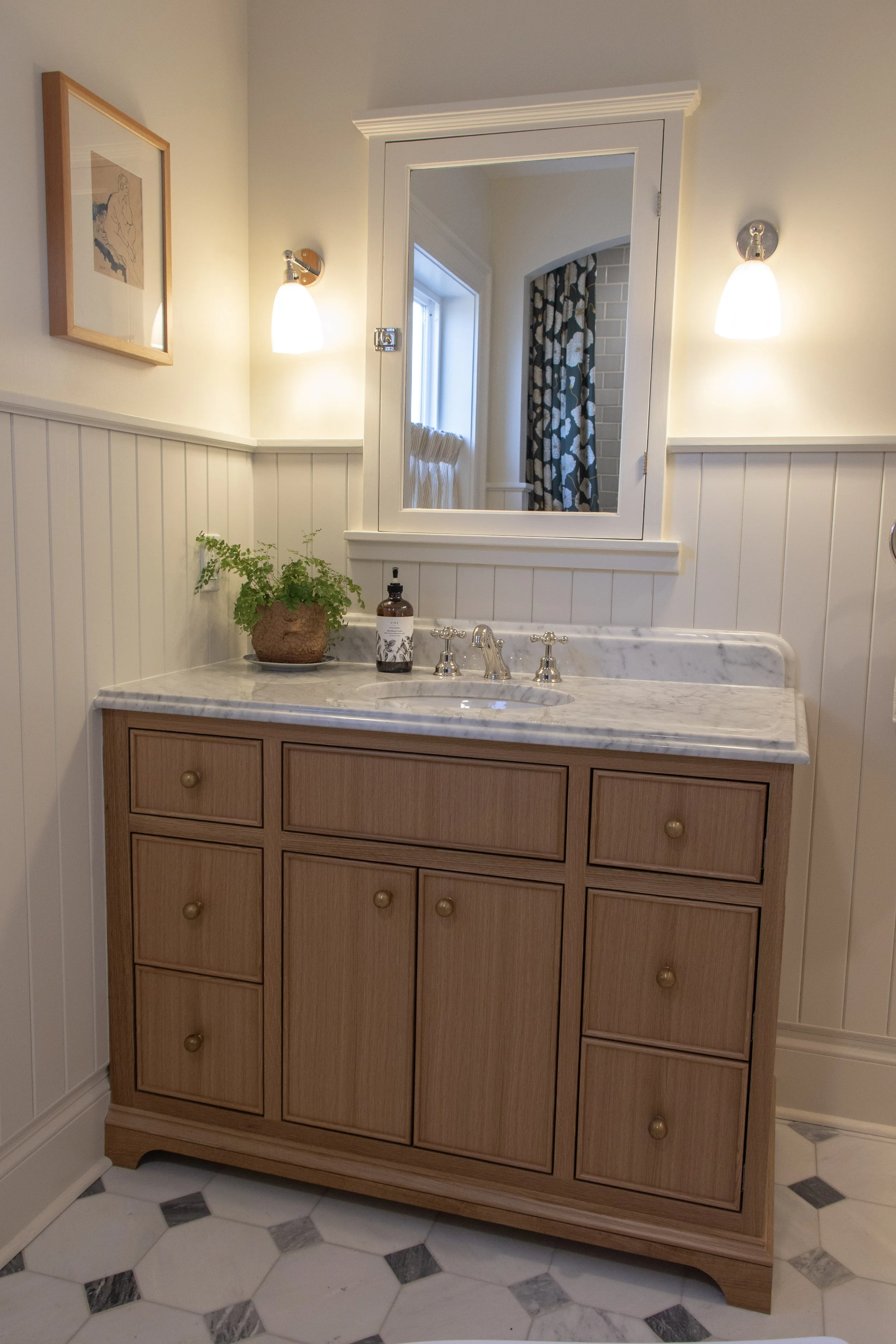 Bathroom vanity with a marble countertop, wooden cabinets, a mirror, wall-mounted lights, potted plant, soap dispenser, and tiled floor.