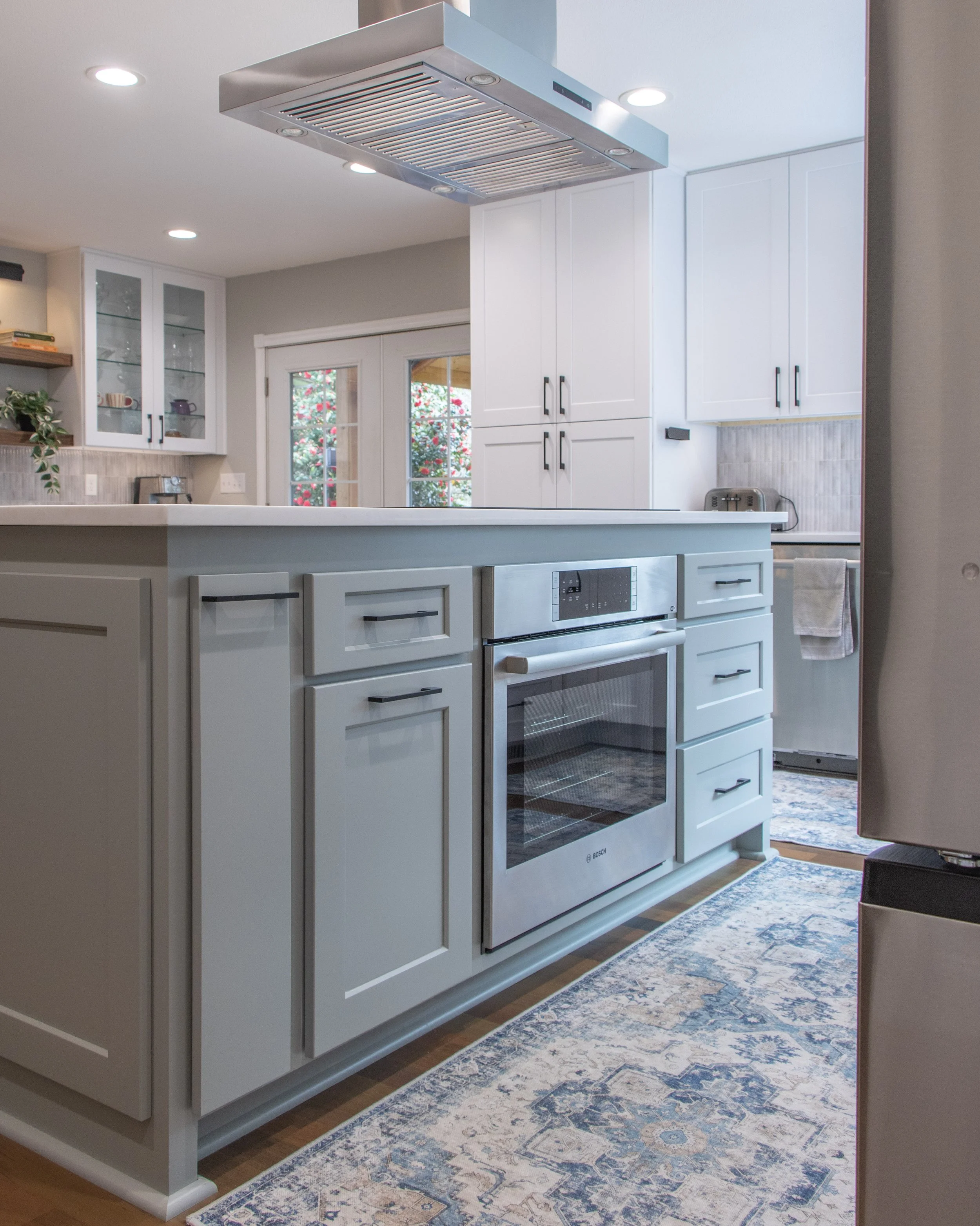 A modern kitchen with white cabinets, a built-in microwave, and a stainless steel oven. There is a kitchen island with a white countertop, and a hanging stainless steel range hood above. The kitchen has recessed lighting, a sliding glass door, and a 