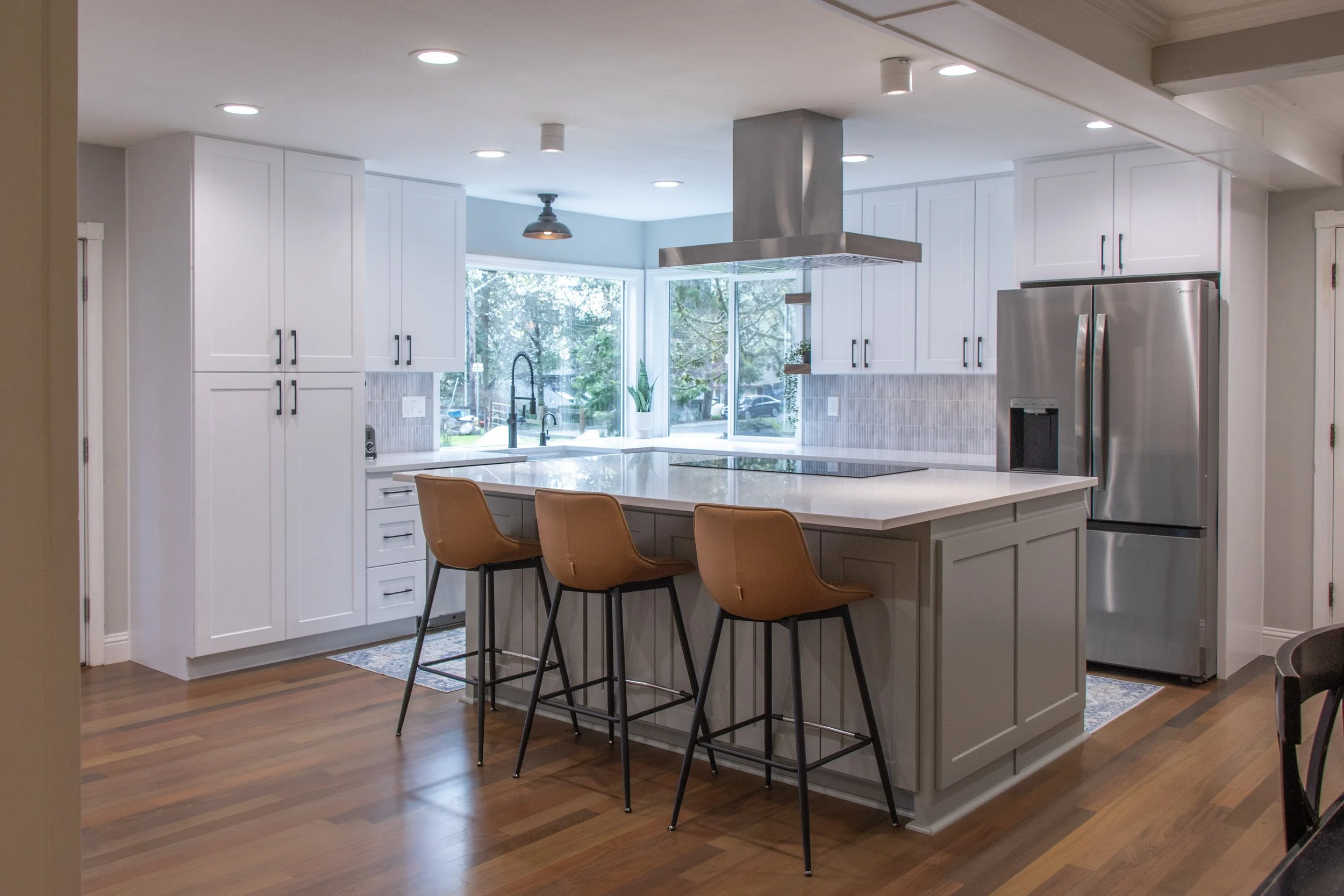 Modern kitchen with white cabinets, a large island with seating, stainless steel refrigerator, large windows with trees outside, and hardwood floors.