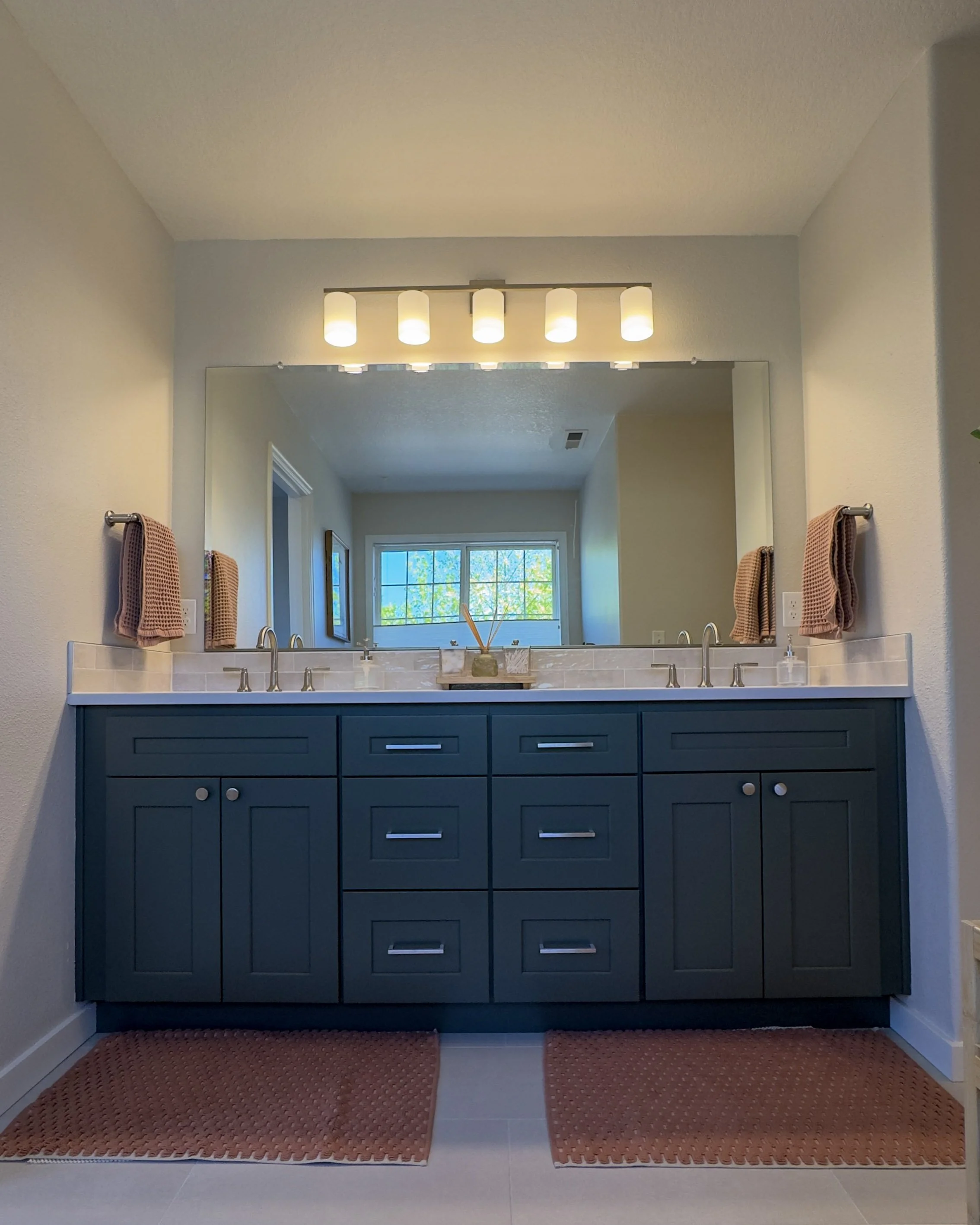 Bathroom with a large mirror above a blue vanity, beige wall-mounted towels, and tiled floors. Overhead light fixture with five cylindrical lights. Window in background with greenery outside.