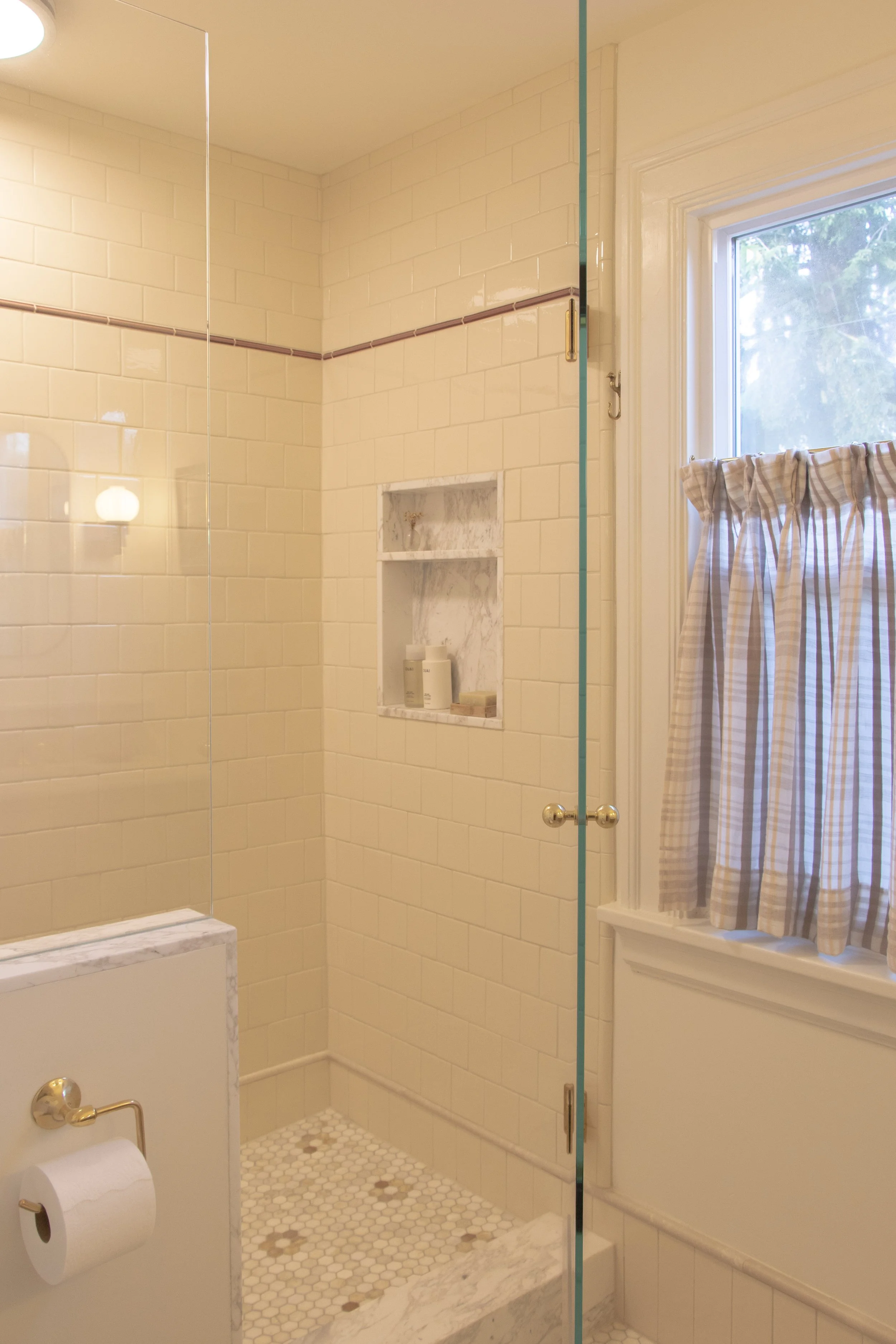 Bathroom shower area with beige tiles, a built-in marble niche, and a window with checkered curtains.