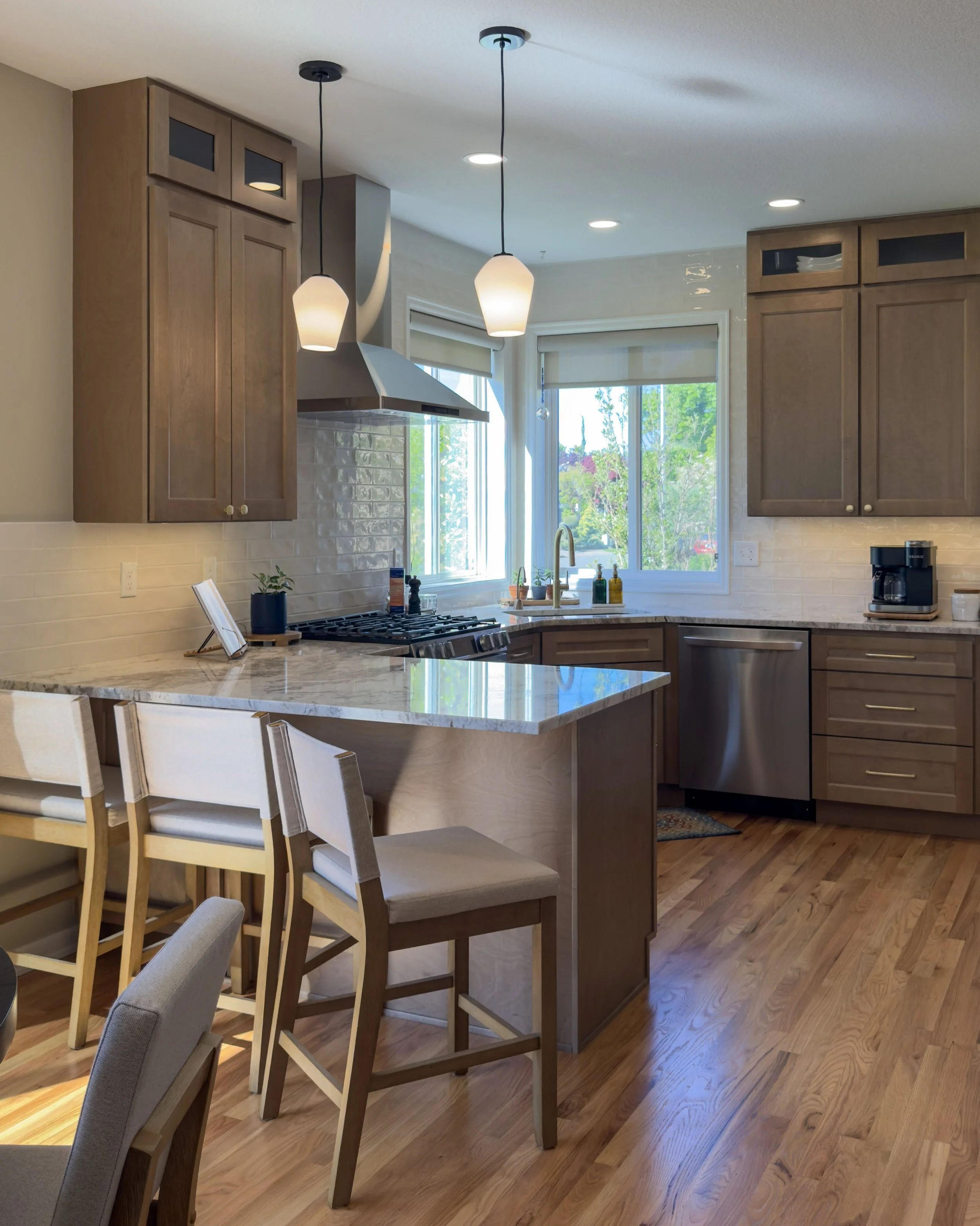 A modern kitchen with wooden cabinets, a breakfast bar with chairs, and a window showing green trees outside. It also features pendant lighting and a marble countertop.