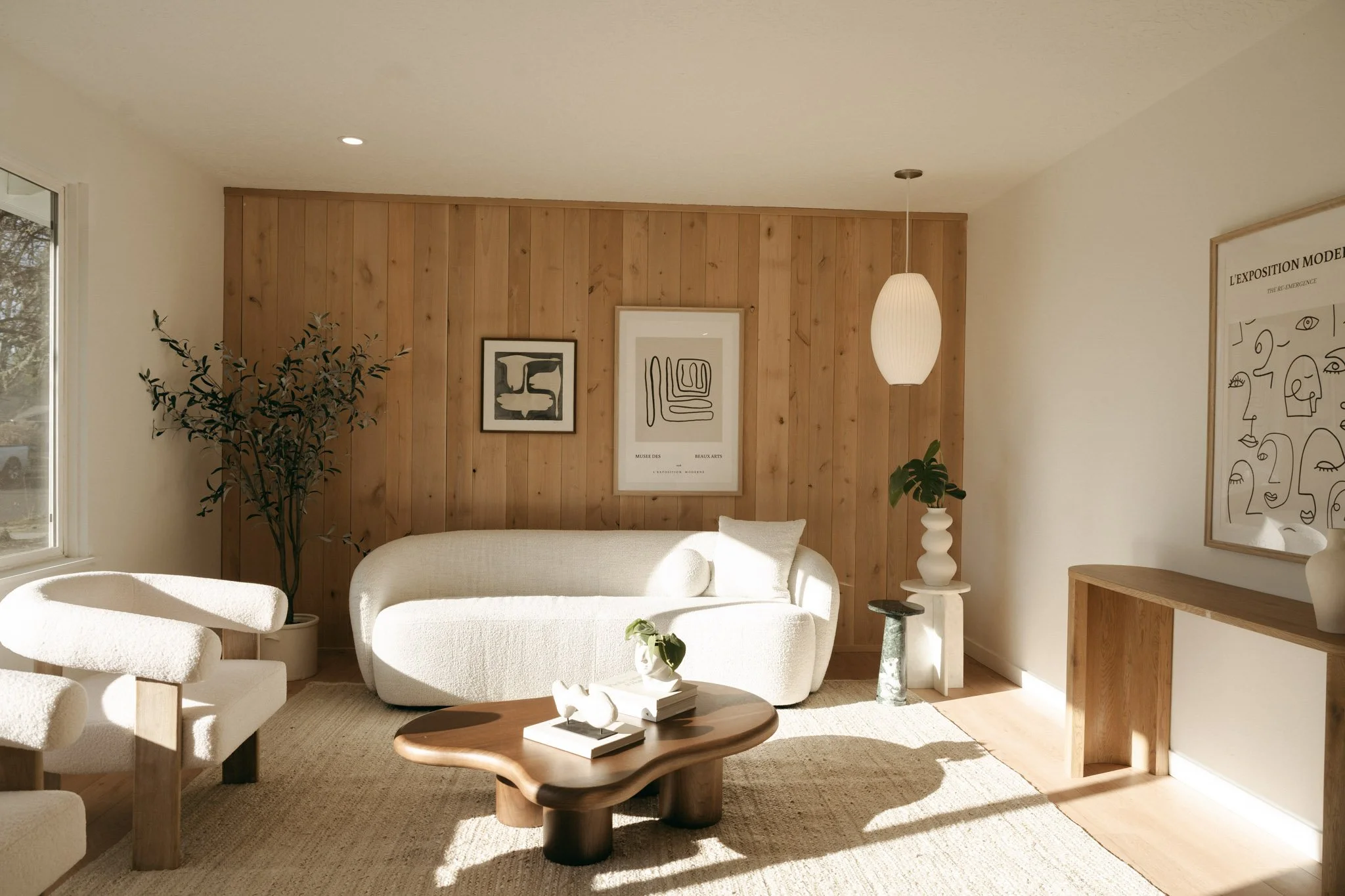 Bright living room with white sofa, armchair, wooden coffee table, wall art, and potted plants, natural light coming through window.