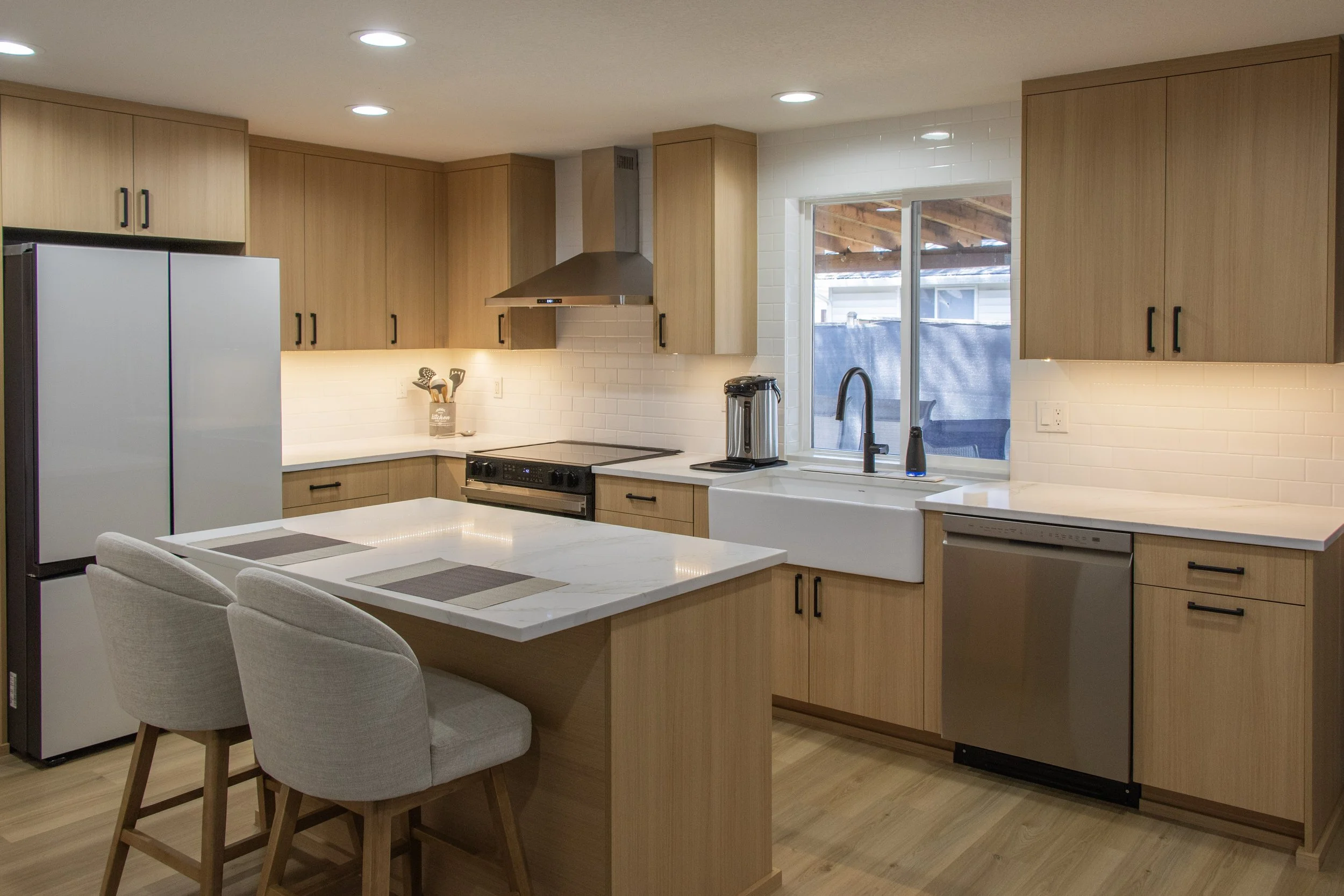 Modern kitchen with wooden cabinets, white subway tile backsplash, stainless steel appliances, a white farmhouse sink, a window, and a white island with chairs.
