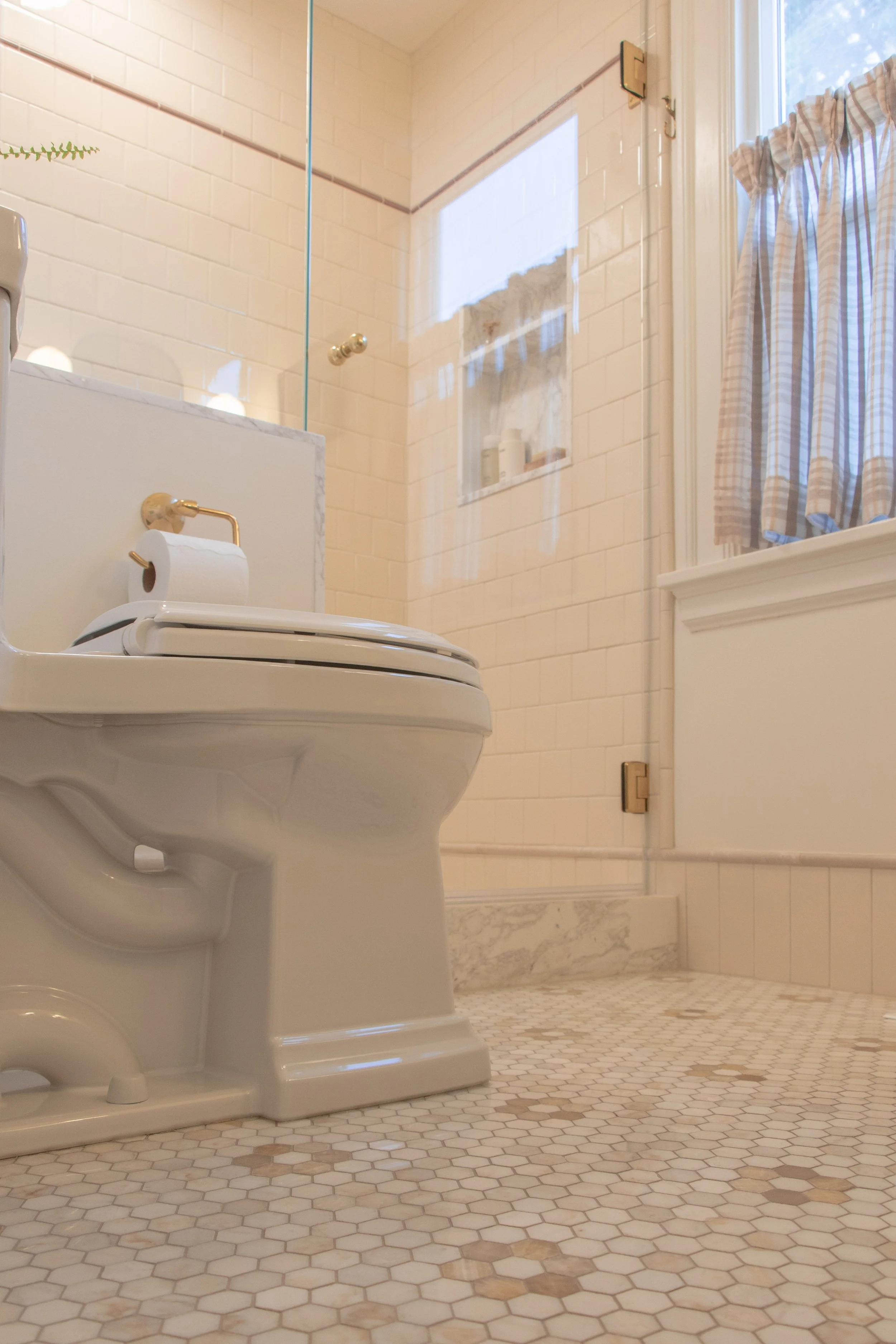 Bathroom with a toilet, a marble ledge, and a shower with a glass door. The bathroom has cream-colored tiles and a window with checkered curtains.