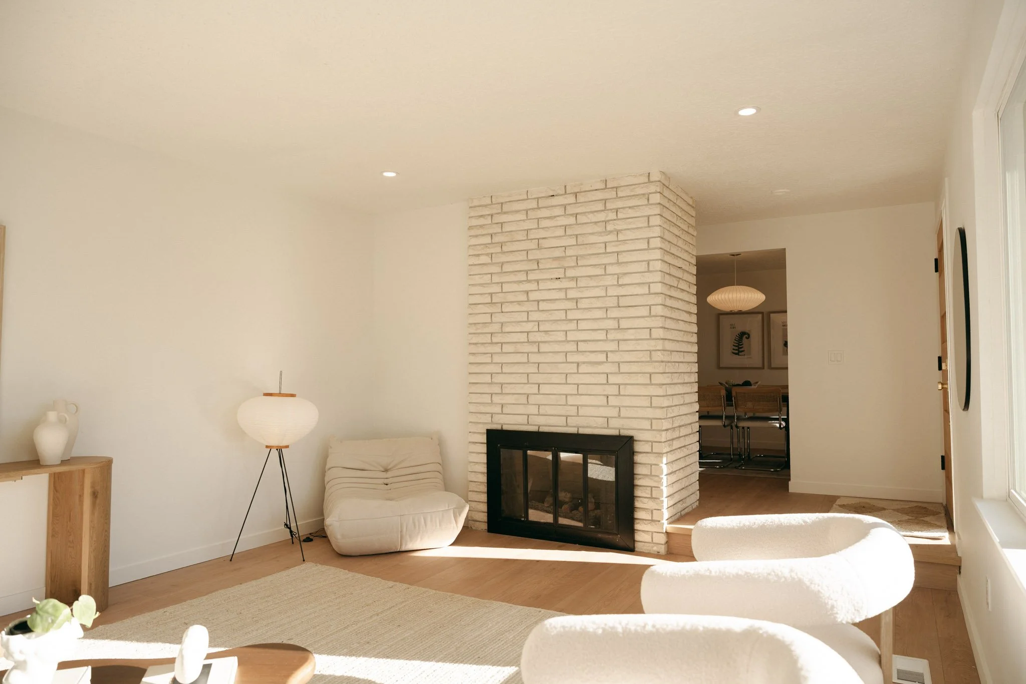 Living room with white walls, a white brick fireplace, white chairs, a wooden side table with vases, a floor lamp, and sunlight streaming in through a window.