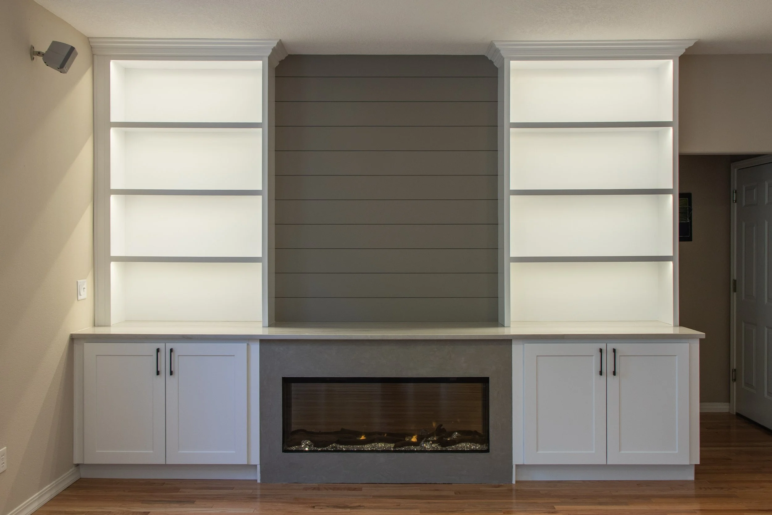 White built-in shelves with lighting, cabinets below, and a fireplace in the center of the lower section, in a room with hardwood floors and beige walls.