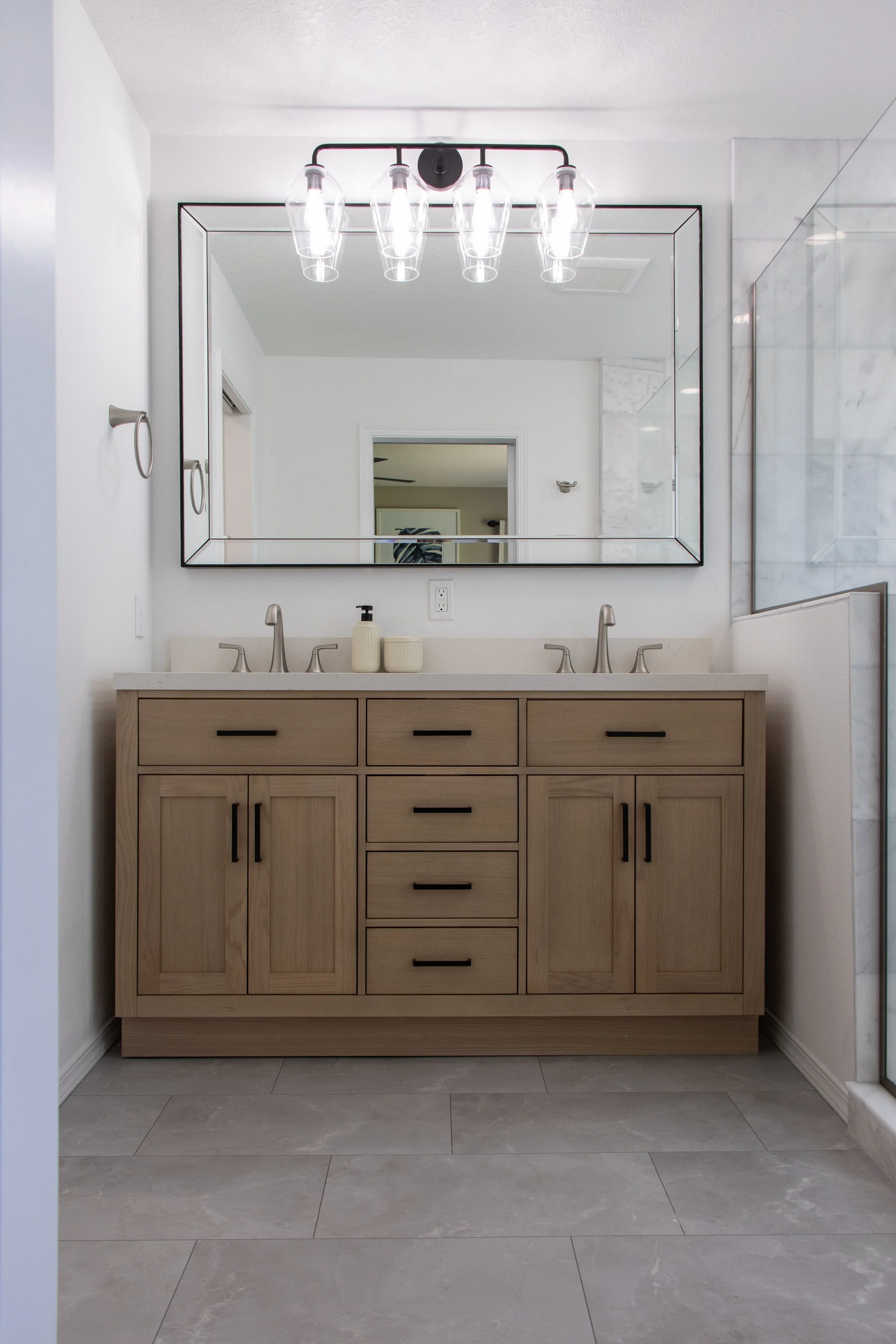 Modern bathroom vanity with a large mirror, double sinks, light-colored wooden cabinet, and beige countertop. There are two soap dispensers and a white electrical outlet visible. The walls are white, and the floor has large gray tiles.