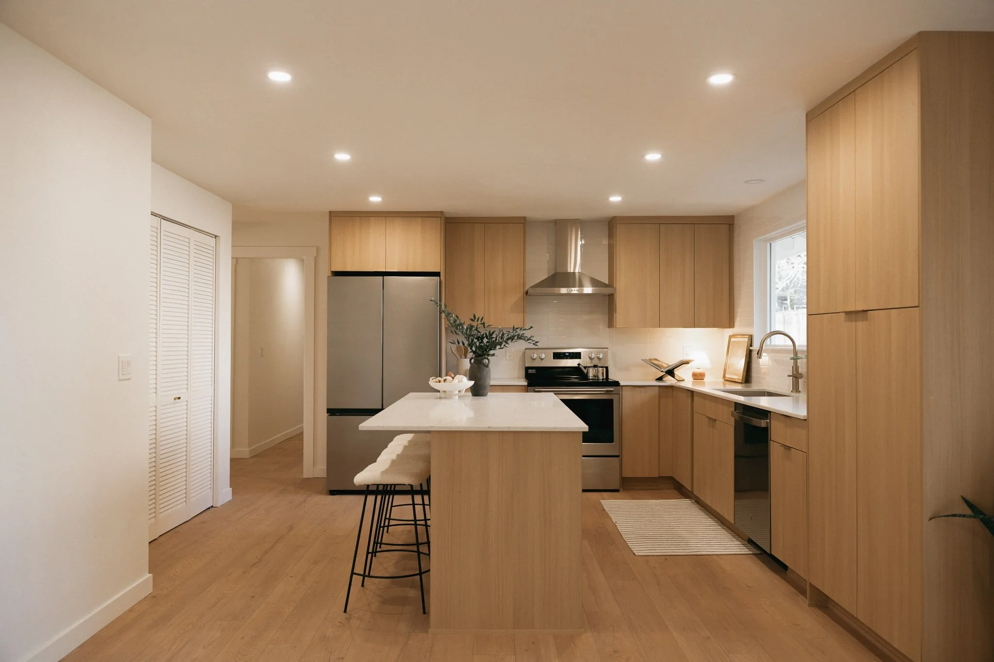Modern kitchen with light wood cabinets, stainless steel appliances, white island with bar stools, and natural light from window.