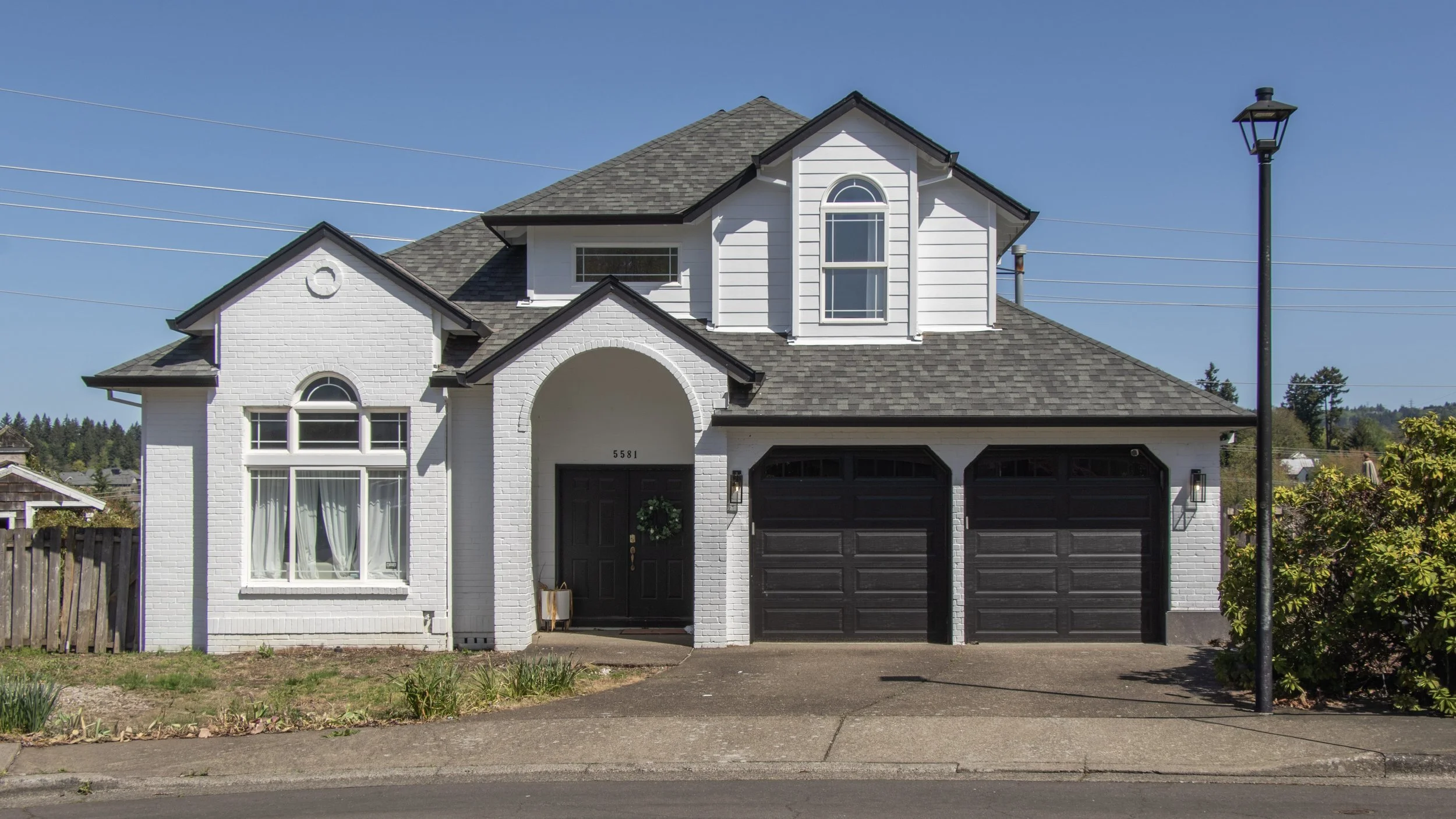A two-story white house with a black garage door, black front door with a wreath, and multiple windows with brown frames, under a blue sky.