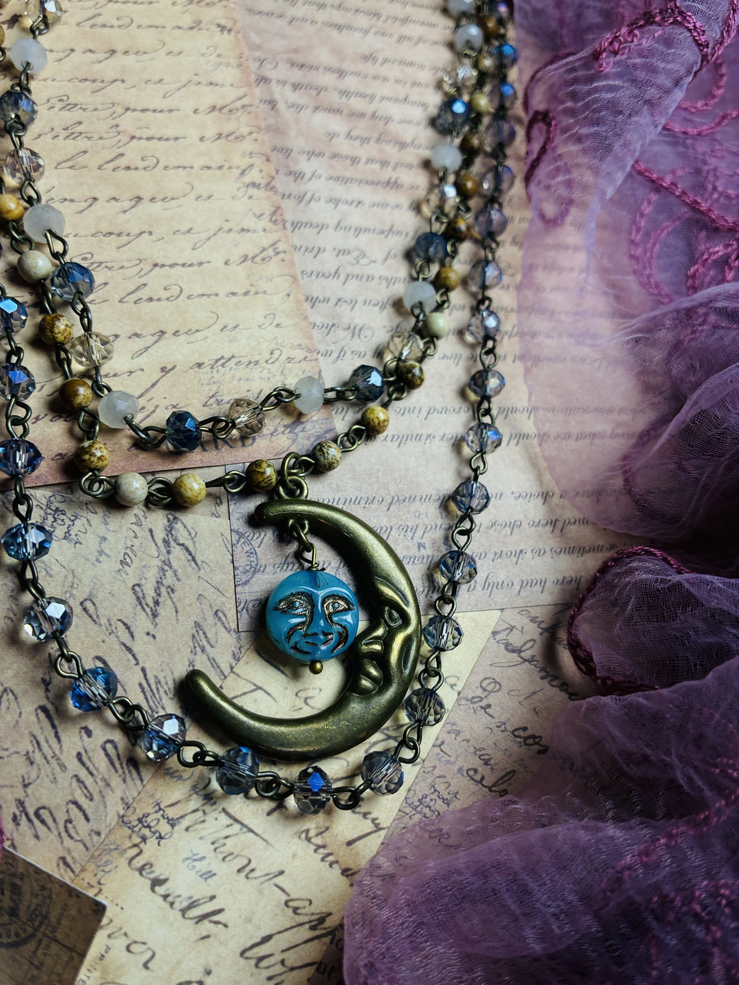 Close-up of the Celestial Moon necklace’s antique-brass crescent pendant, turquoise-blue face charm, and blue crystal beads.