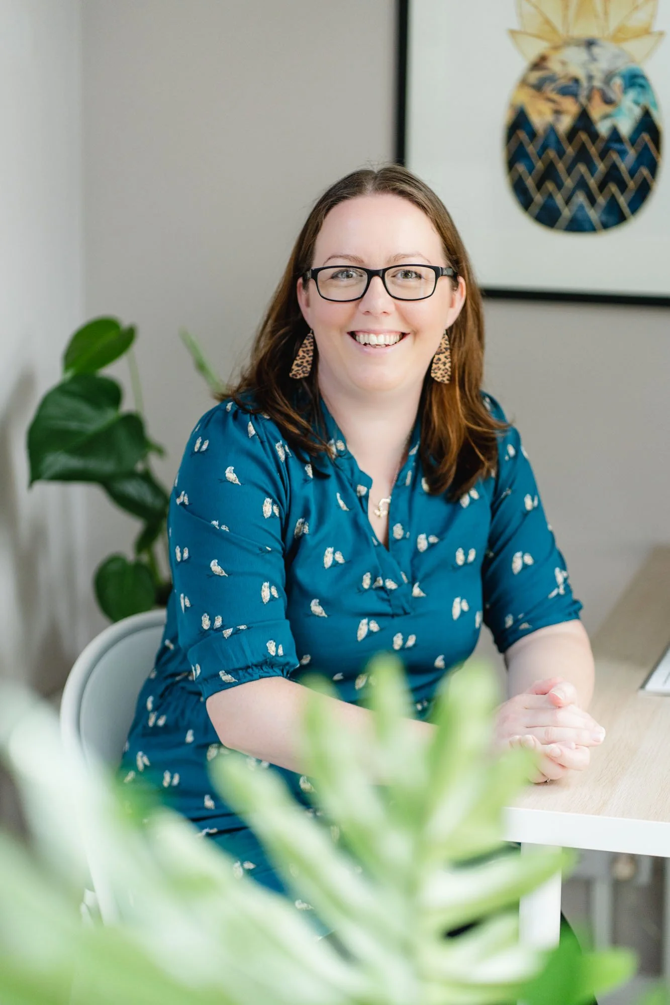 Smiling woman with glasses and leopard print earrings sitting at a desk with houseplants and a pineapple artwork in the background.