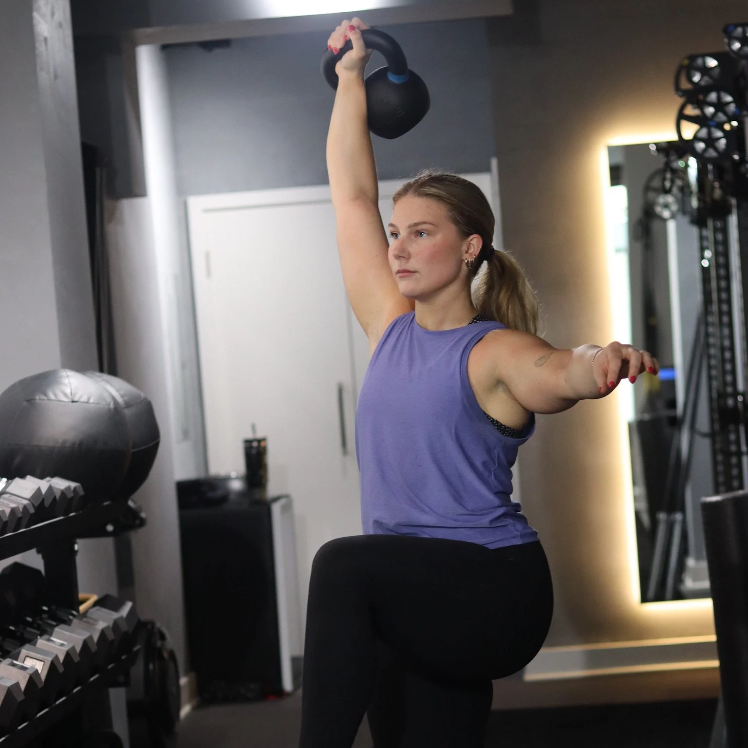 A woman wearing a purple sleeveless top is working out in a gym, lifting a black kettlebell overhead with her right arm, while her left arm is extended outward.