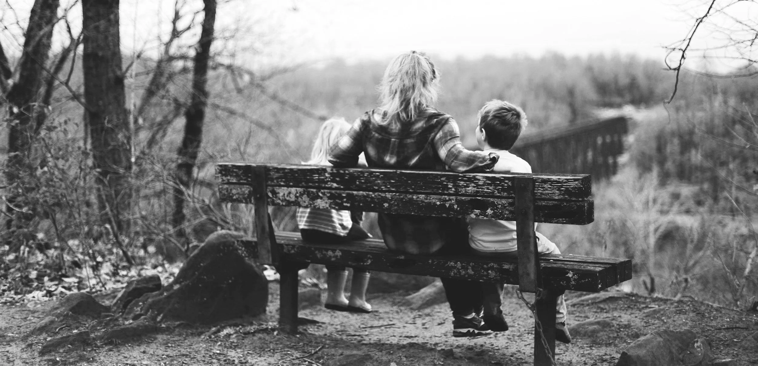 Black and white photo of three people sitting on a bench overlooking a forested valley. Two children sit beside an adult with trees and ridges in the background. The scene is serene and timeless.