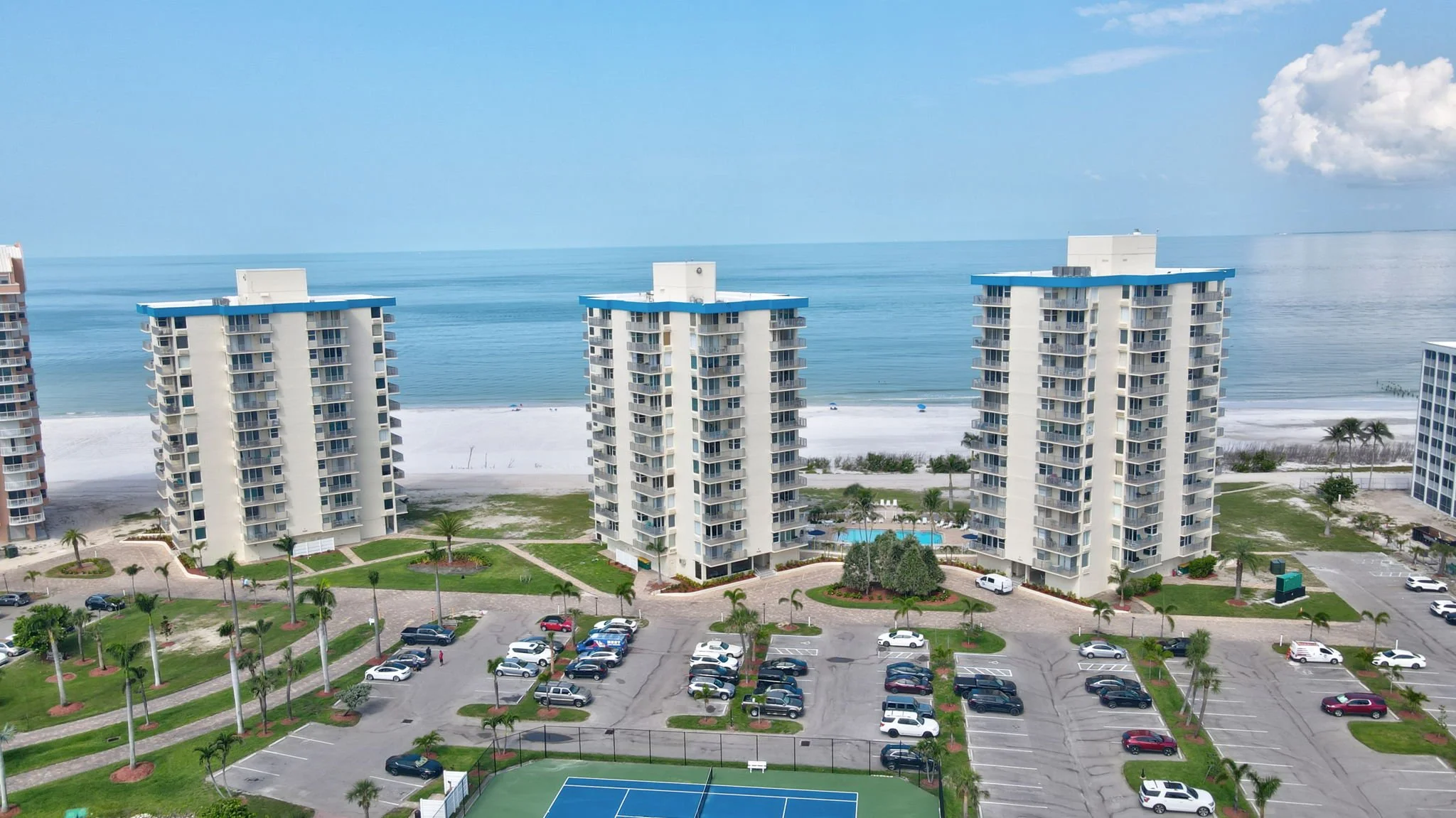 High-rise beachfront resort with three white buildings, swimming pool, parking lot, tennis court, and ocean in the background