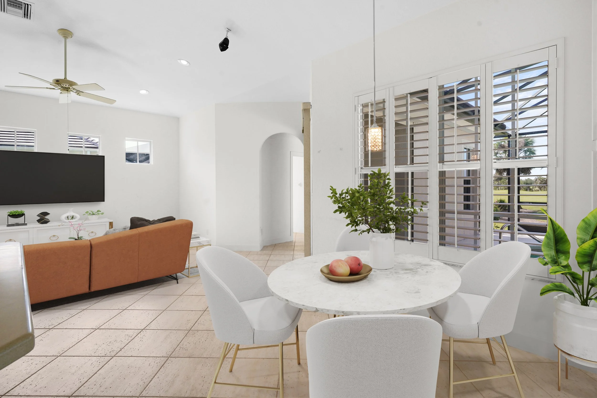 White dining room with a round marble table, four white chairs, potted green plant, and window shutters; open plan living area with a tan sofa, TV, ceiling fan, and large windows.