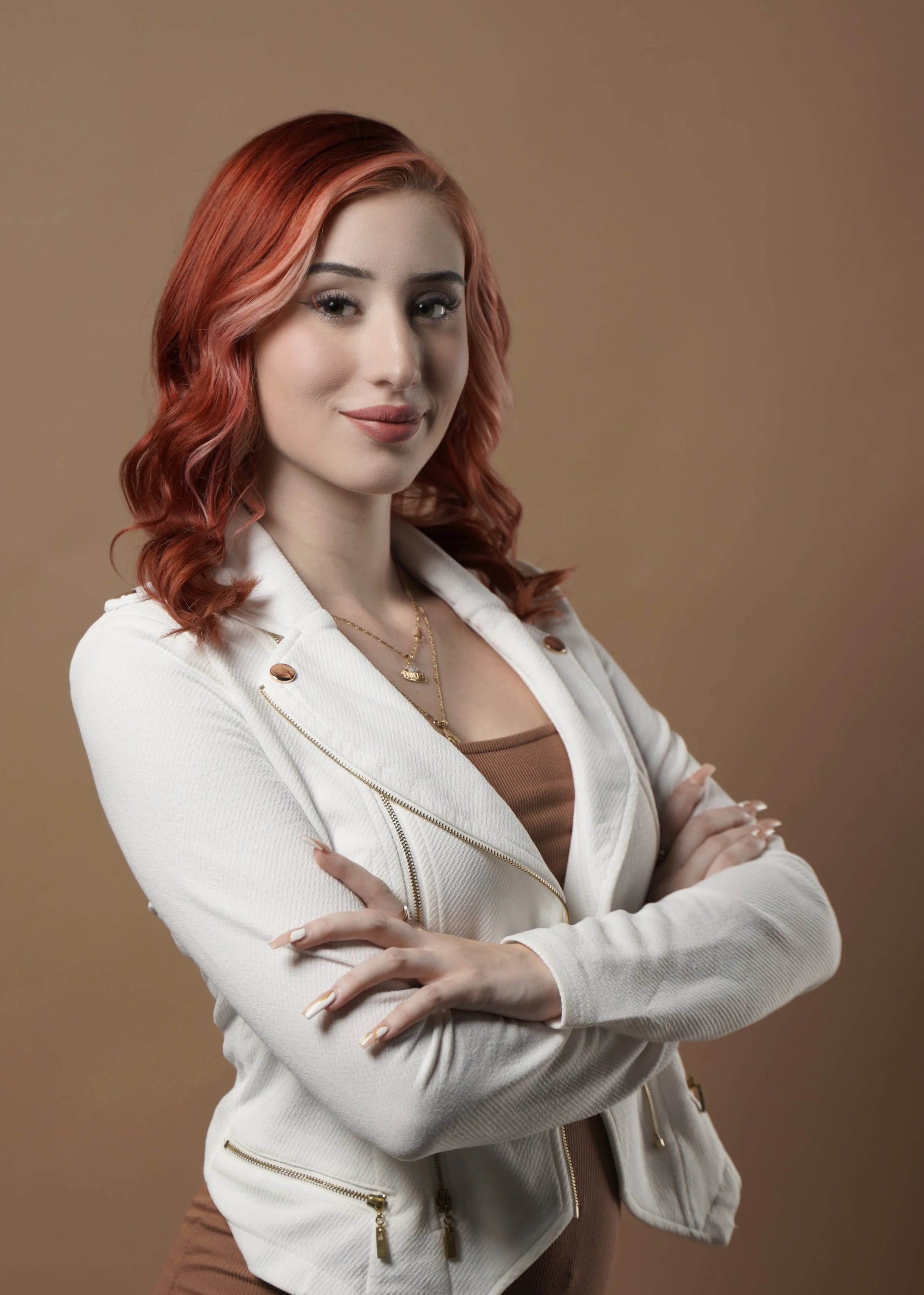 A young woman with red wavy hair, wearing a white jacket over a beige top, crosses her arms and looks at the camera with a slight smile
