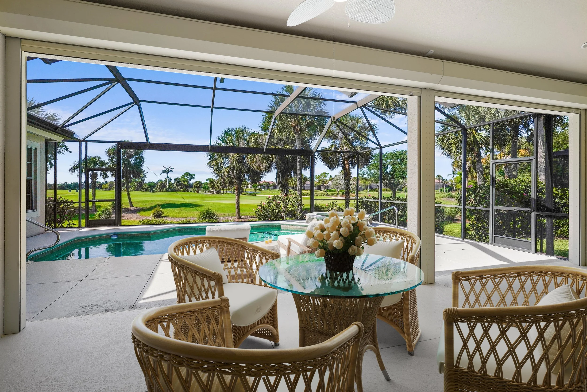 View from inside a screened porch featuring a round glass-top table with a flower arrangement, surrounded by wicker chairs, overlooking a backyard with a swimming pool, palm trees, and a green lawn.