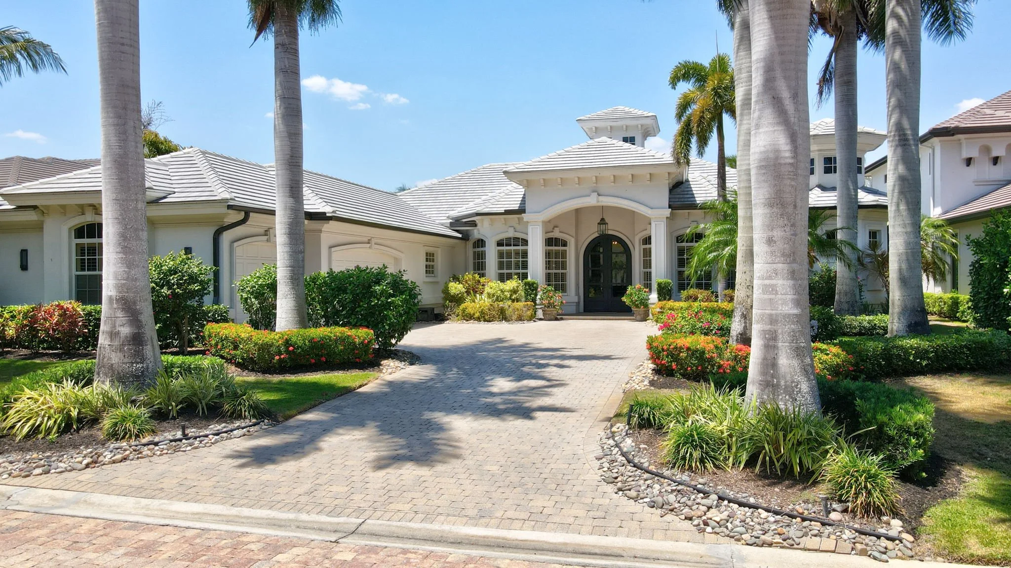 Front view of a large, elegant house with white exterior and gray roof, surrounded by tall palm trees, colorful flowers, and well-maintained landscaping on an sunny day.