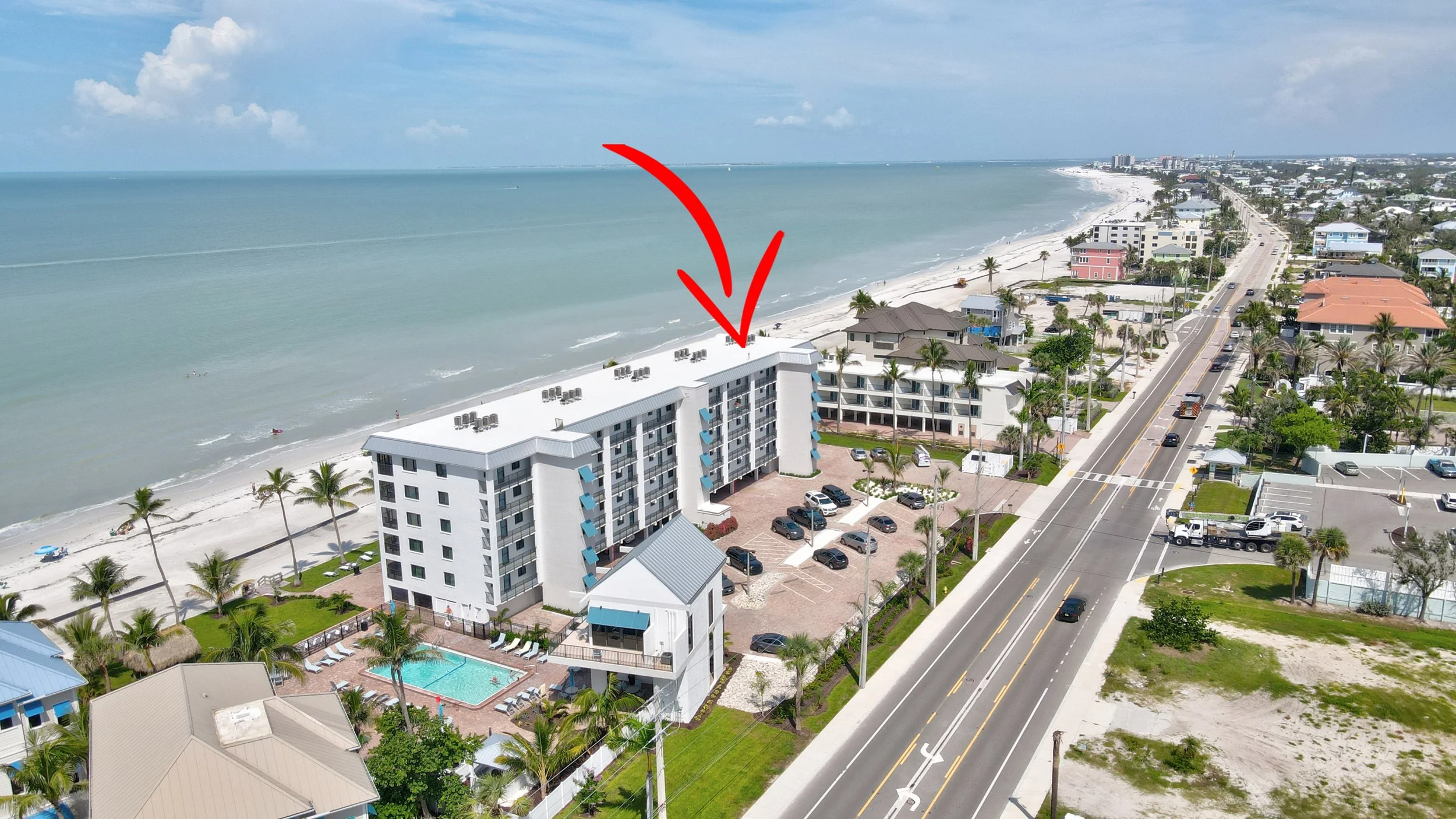 Aerial view of a beachfront hotel with a large white building, parking lot, swimming pool, and palm trees, located along a coastal road with the ocean in the background.