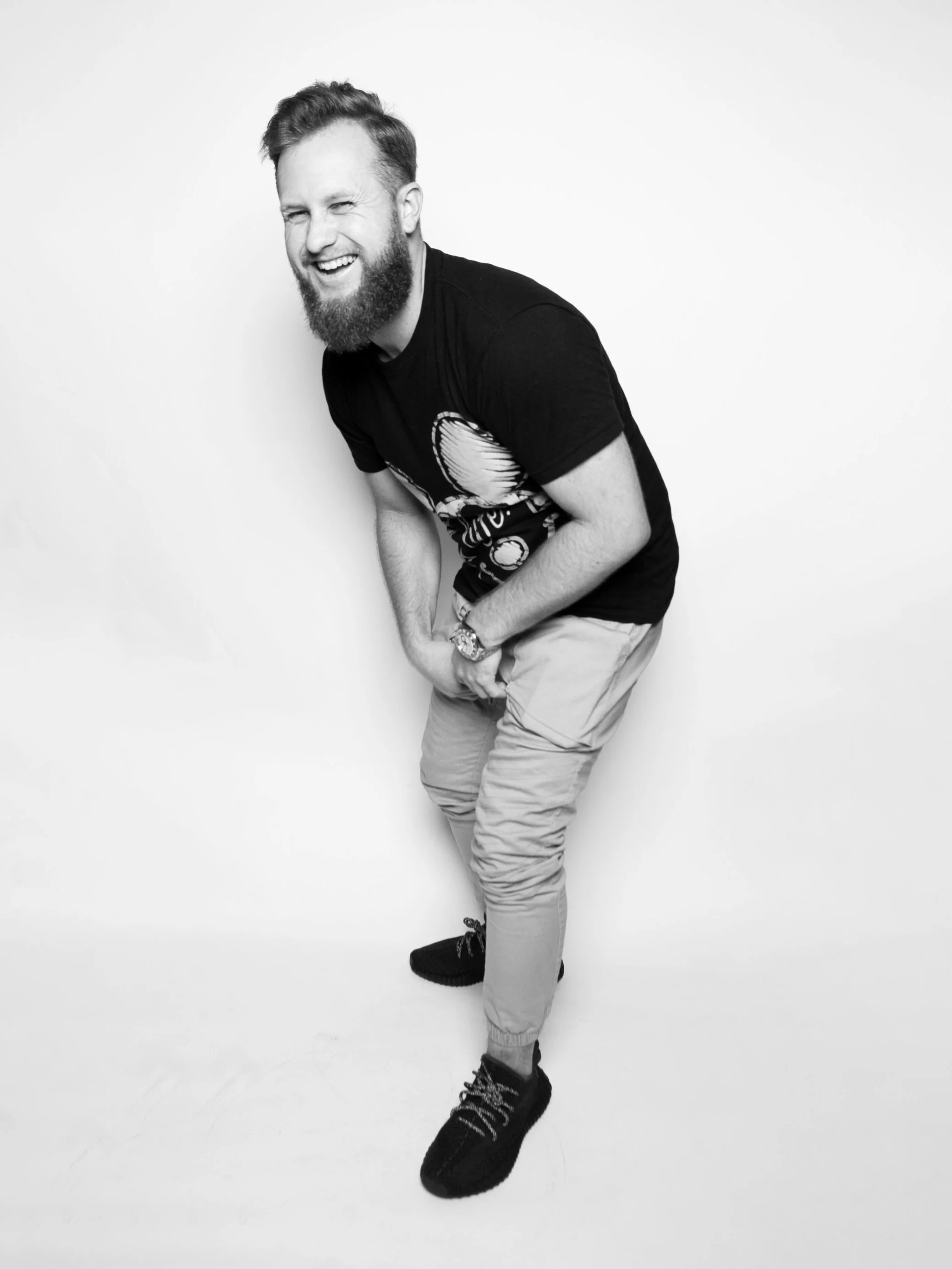 A young man with a beard laughing and leaning forward against a white background, wearing a black t-shirt, light-colored pants, and black sneakers.