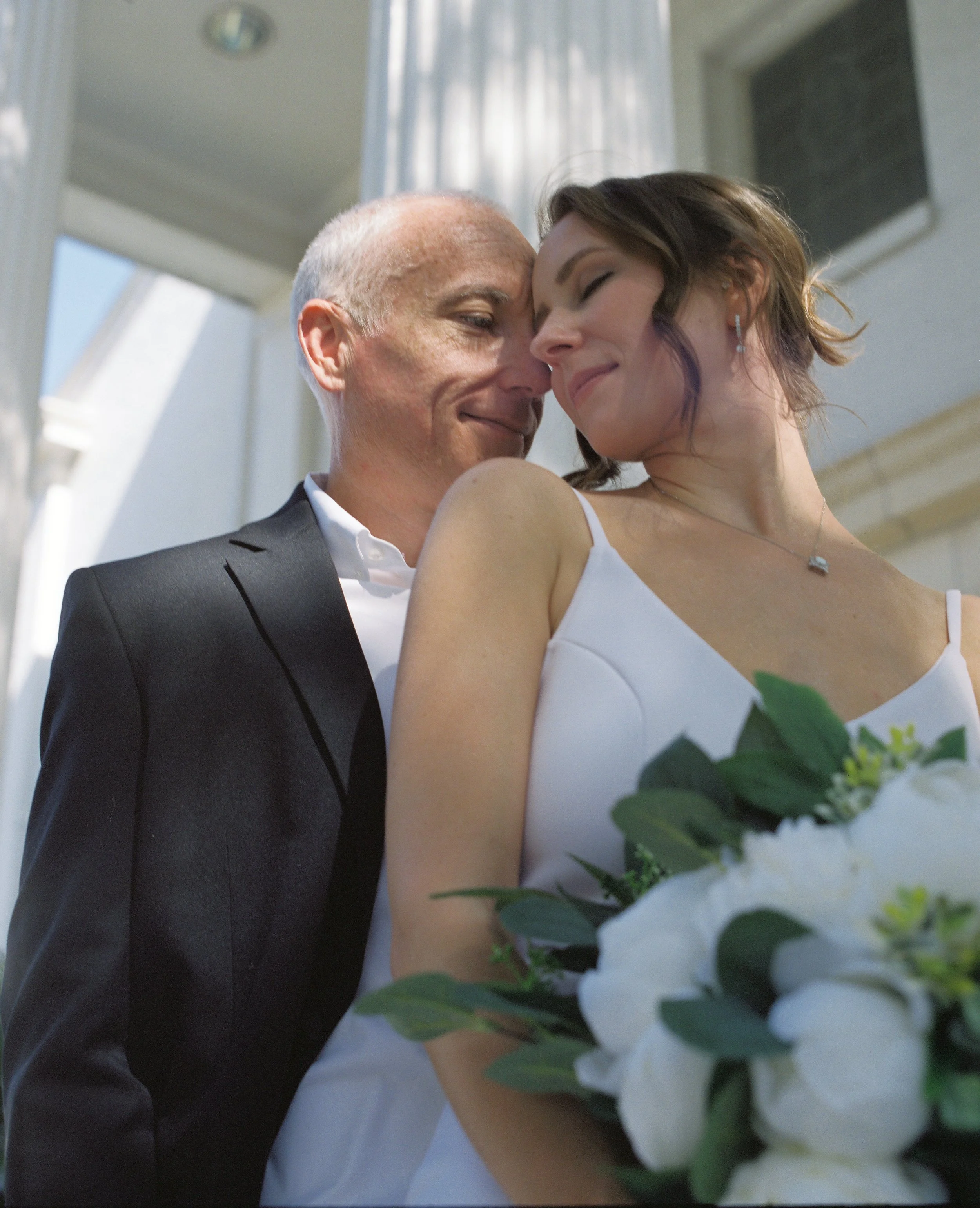 A couple dressed in wedding attire sharing an intimate moment, with the bride holding a bouquet of white flowers and greenery.