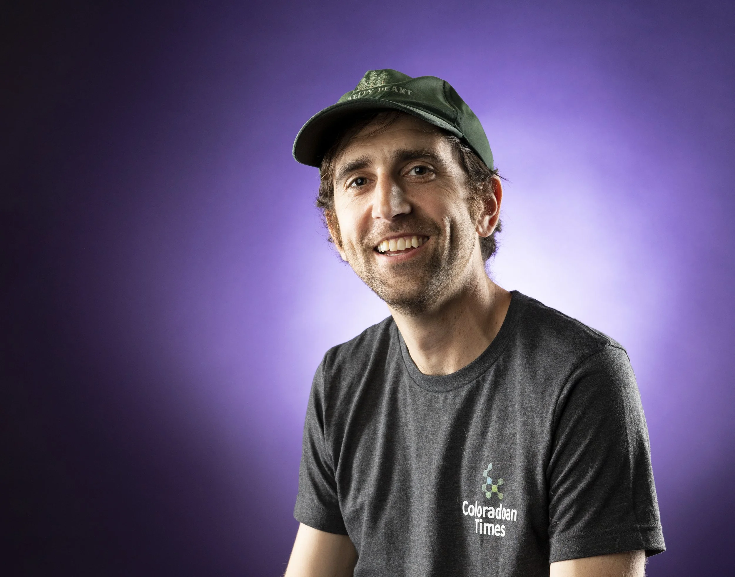 A smiling man wearing a dark gray t-shirt with 'Coloradoan Times' logo and a green cap, posing against a purple gradient background.