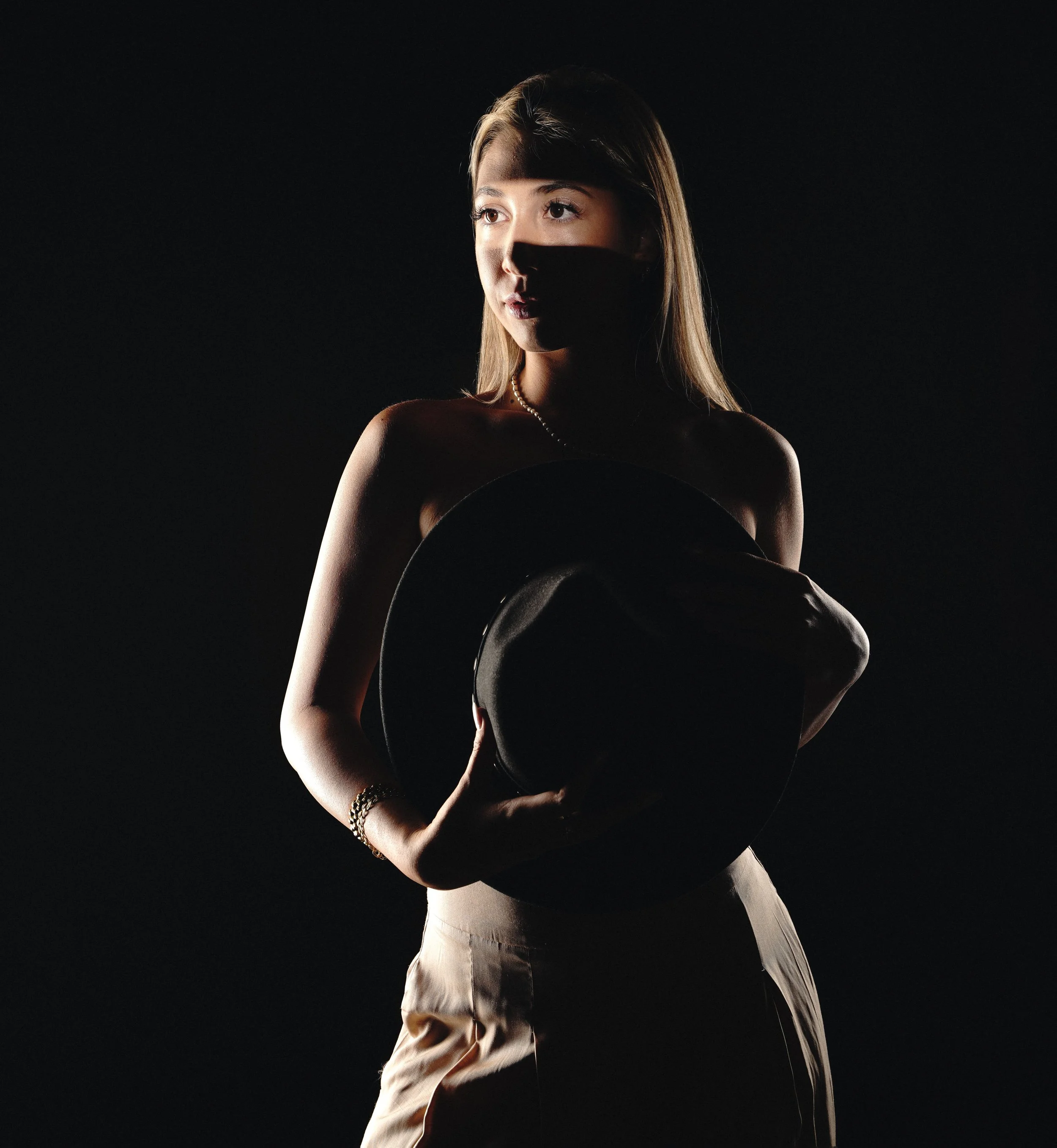 A woman with brown hair and a pearl necklace holding a black hat and a black circular object, standing against a dark background with soft lighting highlighting her face and shoulders.