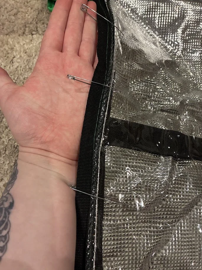 Close-up of a person's hand with a tattooed wrist, holding a reflective silver insulated material secured with safety pins to a black fabric edge.