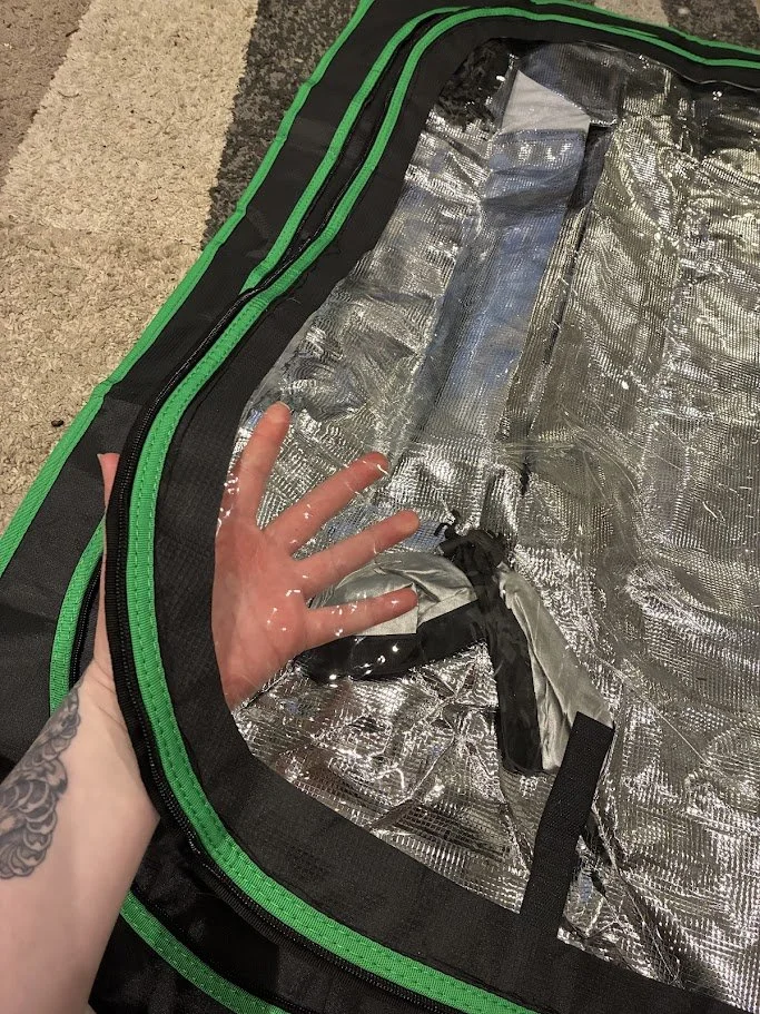 A person's hand is inside a silver, reflective grow tent with an open zipper, showing the interior fabric and black straps.