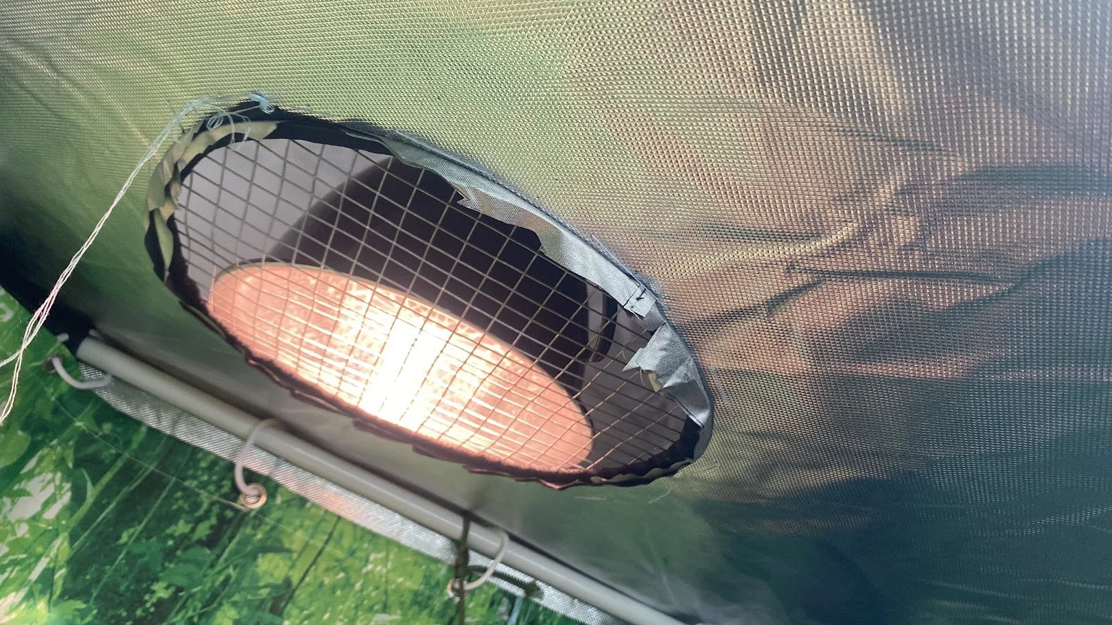 View of an electric heater or heat lamp installed in a fabric or mesh enclosure, with a grid covering the heating element, mounted on a support structure, and blurry green vegetation in the background.
