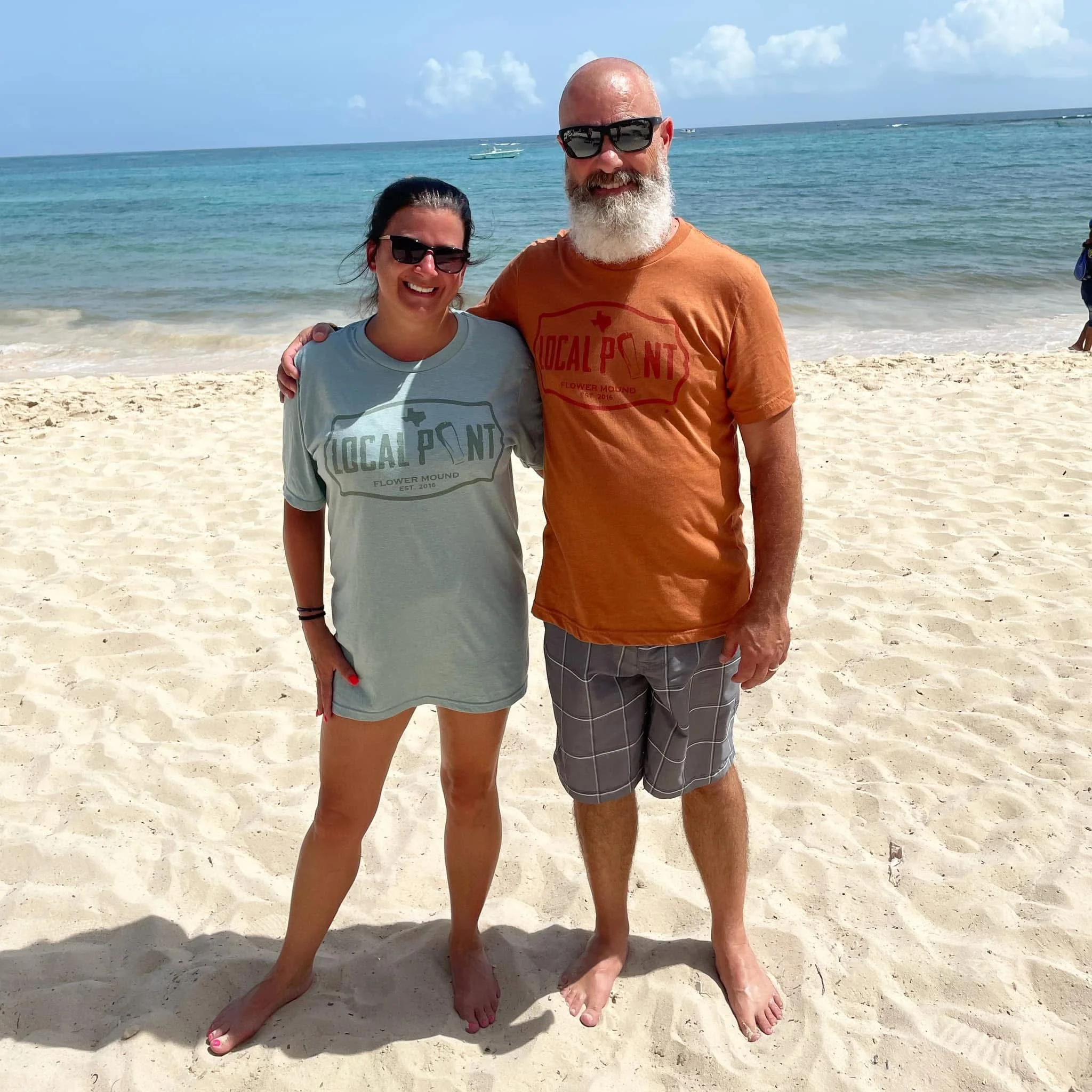 A woman and a man standing on a sandy beach with the ocean and boats in the background.