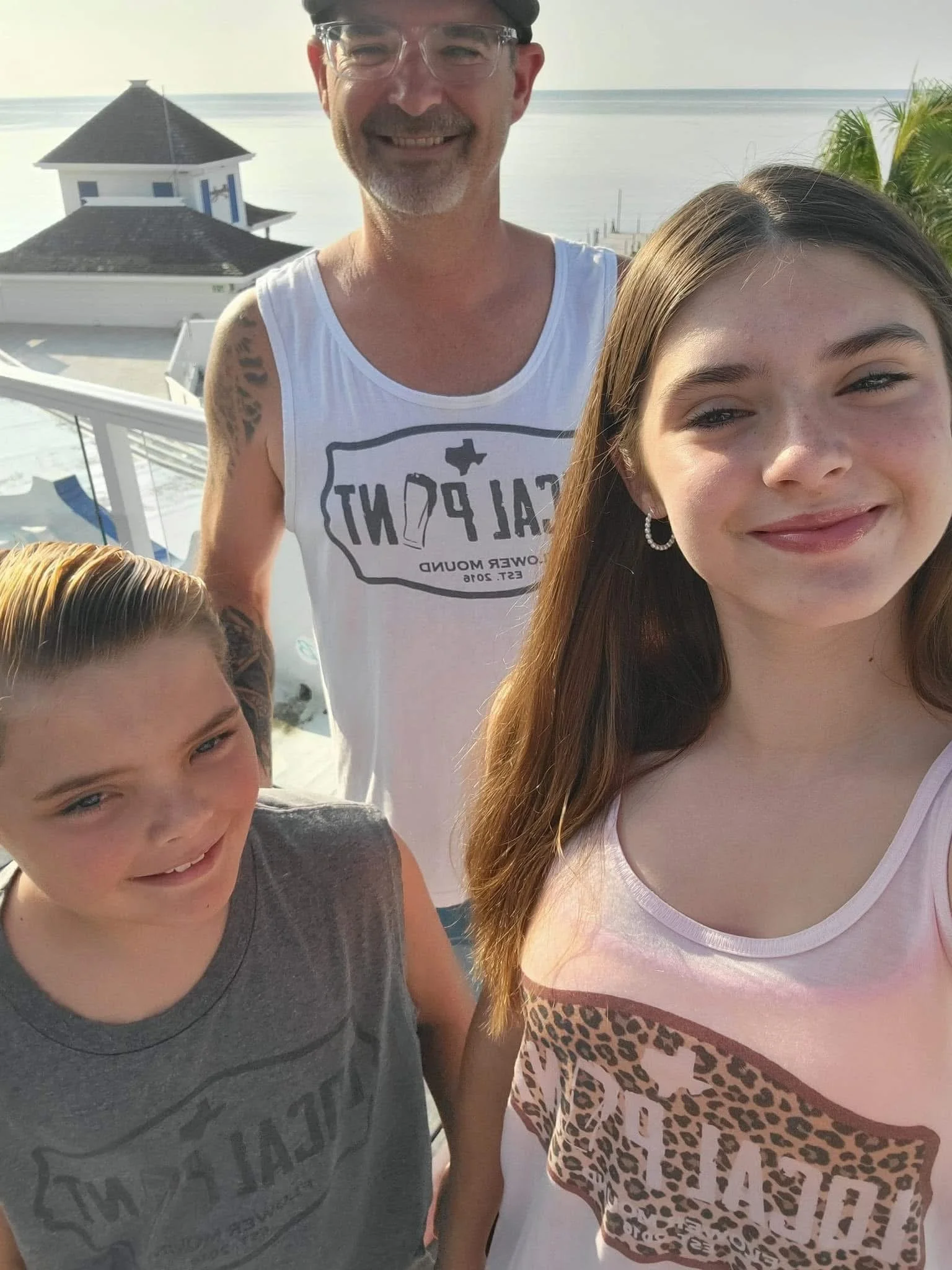 A man and two children taking a selfie on a balcony near the water. The man is smiling, wearing a white tank top and glasses. The girl has long brown hair, is smiling, and wearing a light pink tank top with leopard print. The boy has short blond hair